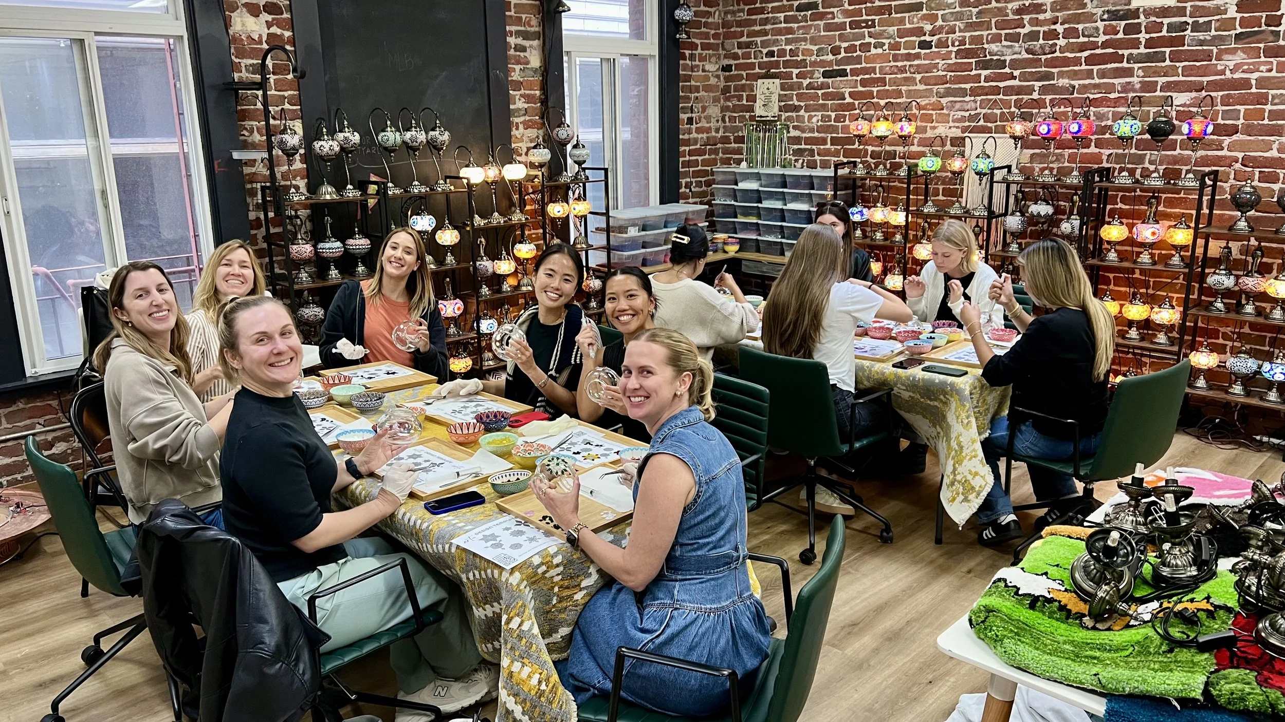 Group of women sitting around a table at a craft workshop, smiling at the camera, with colorful lamps and decorative glass objects in the background.