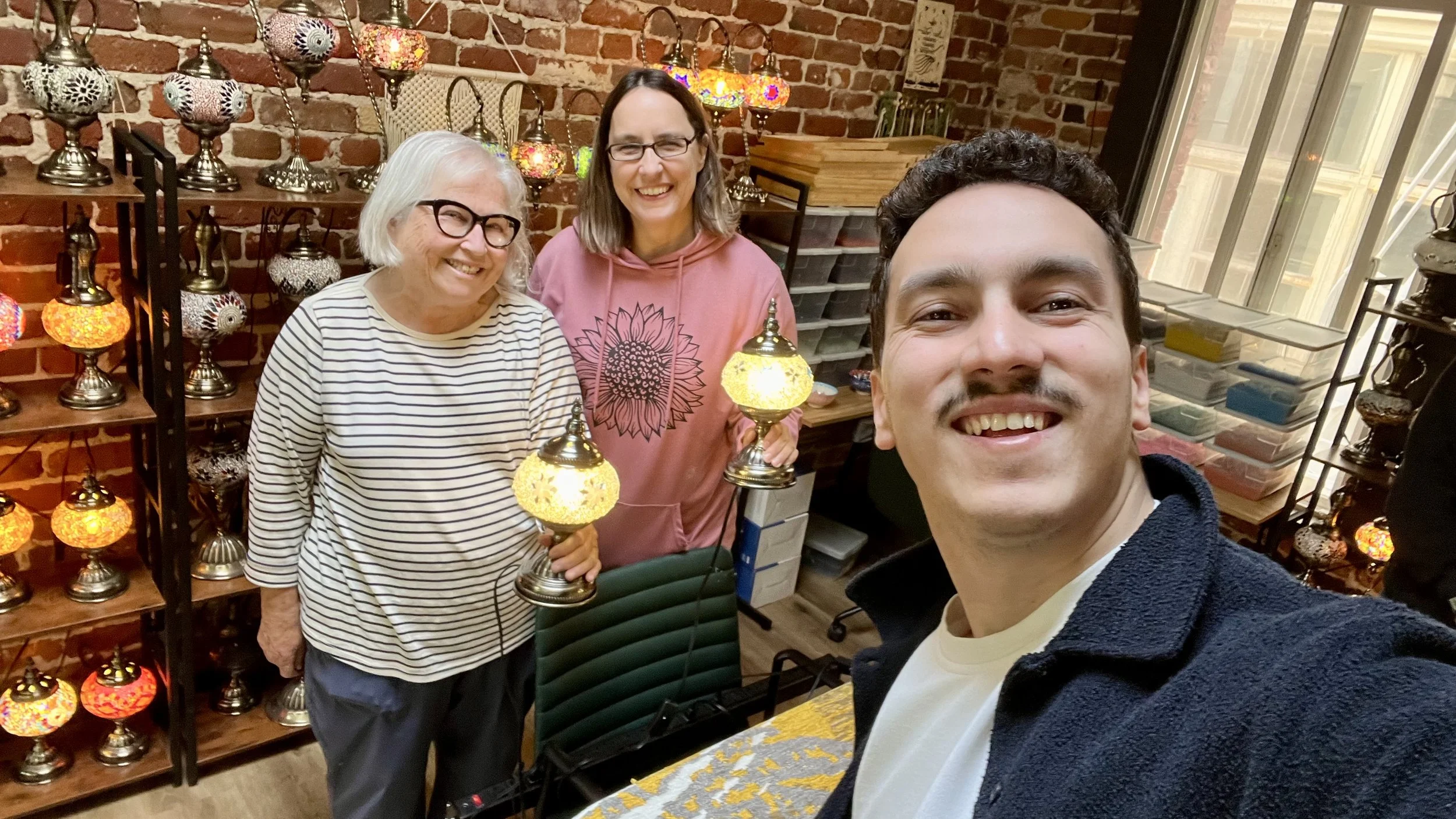 A selfie of three smiling people in a lamp shop, with shelves of colorful mosaic lamps, brick walls, and a large window in the background.