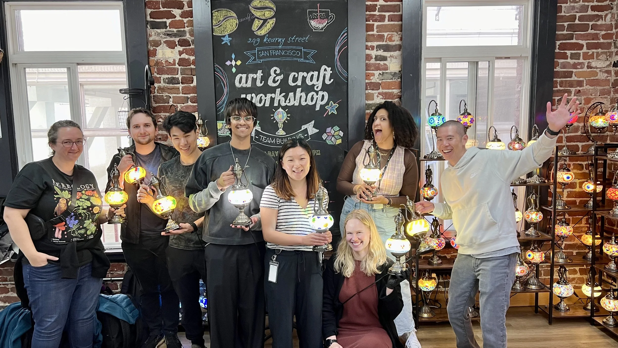 Group of eight people standing and sitting inside a brick-walled art and craft workshop, holding colorful glass lamps, with a blackboard behind them advertising the workshop at 209 Kearny Street in San Francisco.