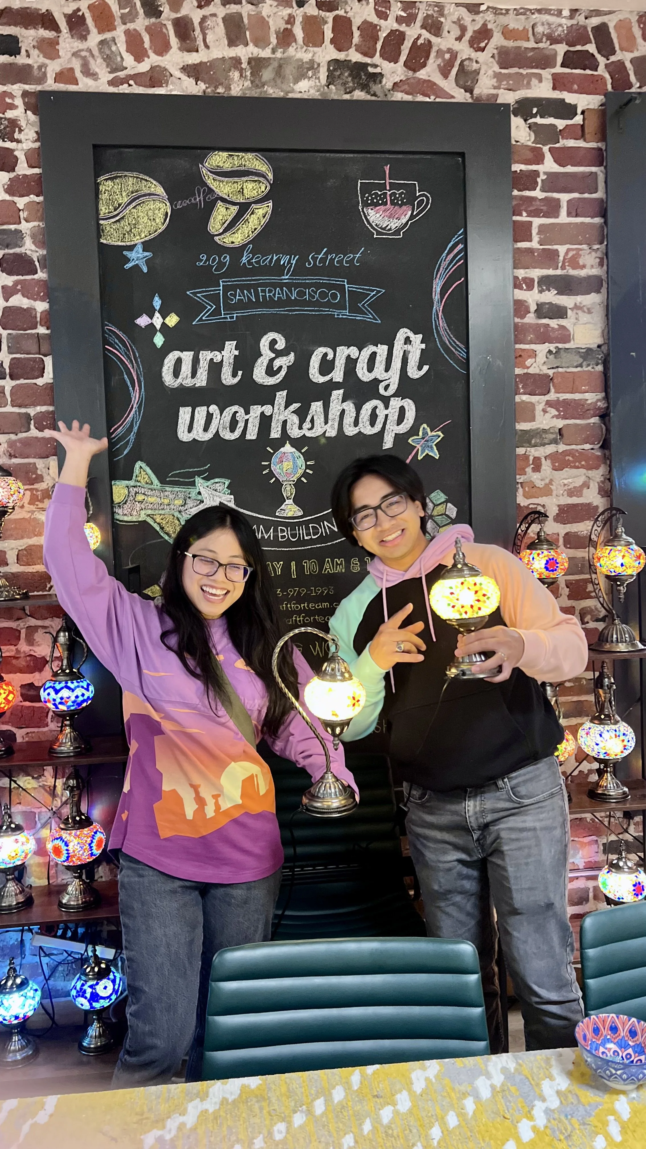 Two people smiling and holding colorful Turkish lamps during an art and craft workshop in a room with a brick wall and a blackboard sign.