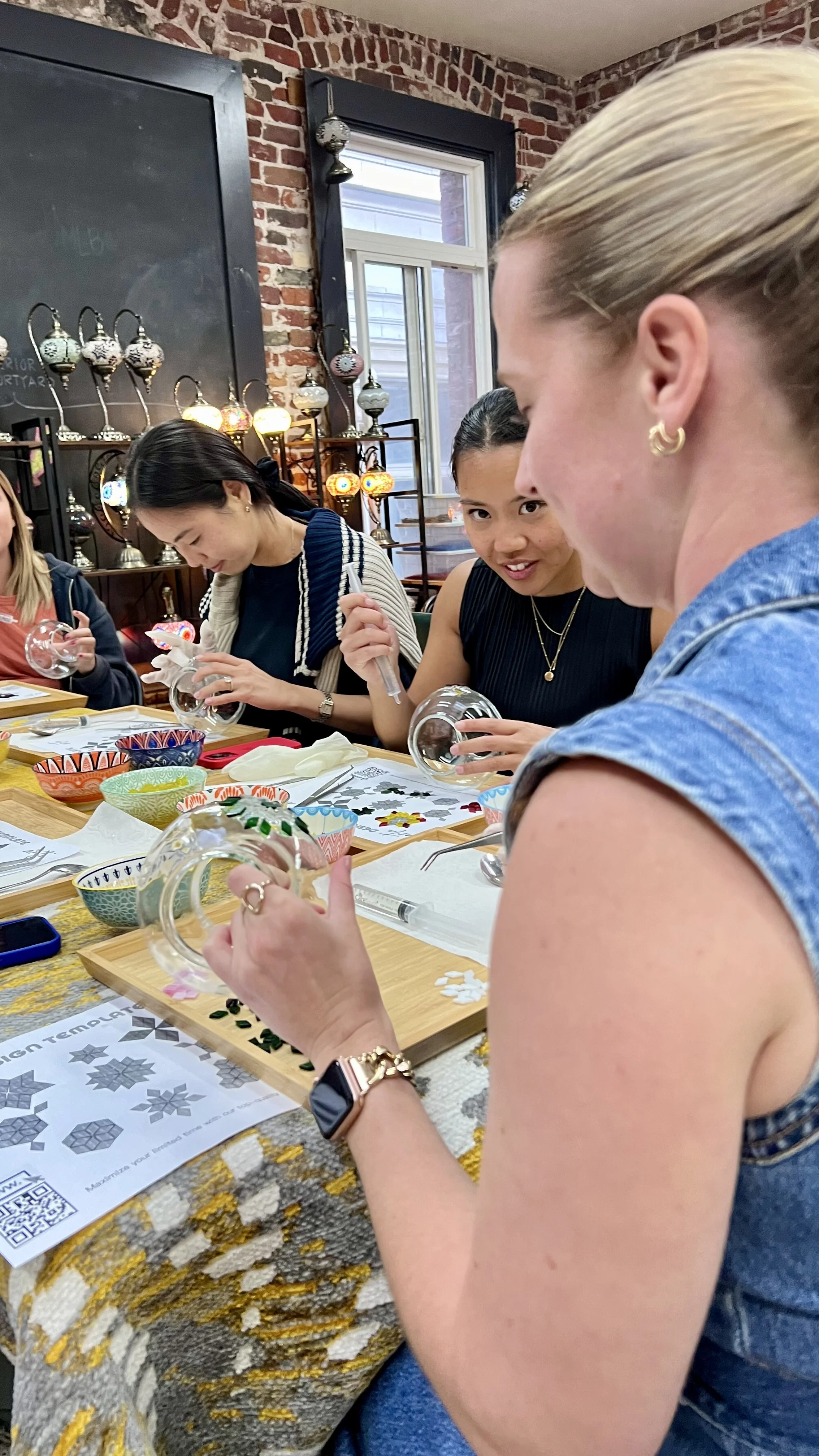 Group of women participating in a glass painting craft workshop in a cozy, brick-walled room with colorful bowls and tools on the table.