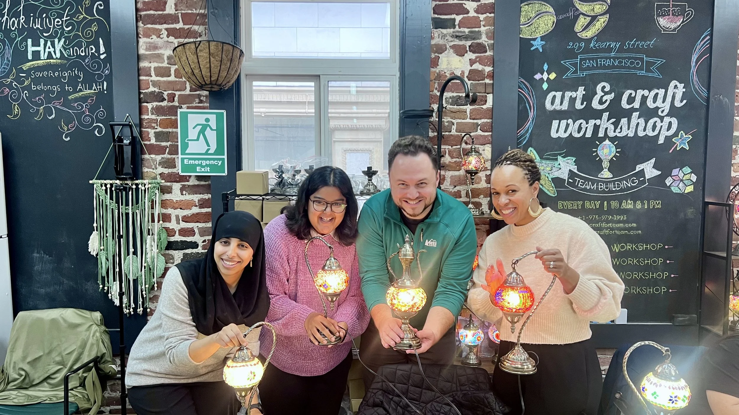Four people smiling and holding colorful mosaic lamps in an art and craft workshop, with a blackboard sign behind them displaying workshop details, in a cozy brick-walled room.