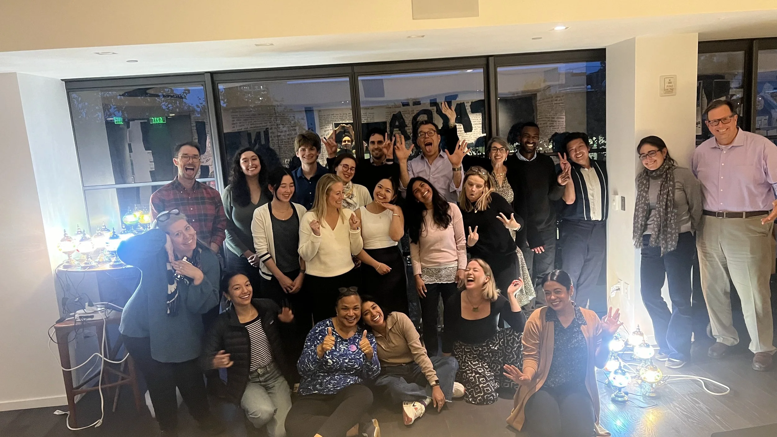 Group of people celebrating indoors with smiles and peace signs, standing and sitting, in front of glass doors, near decorative lamps.
