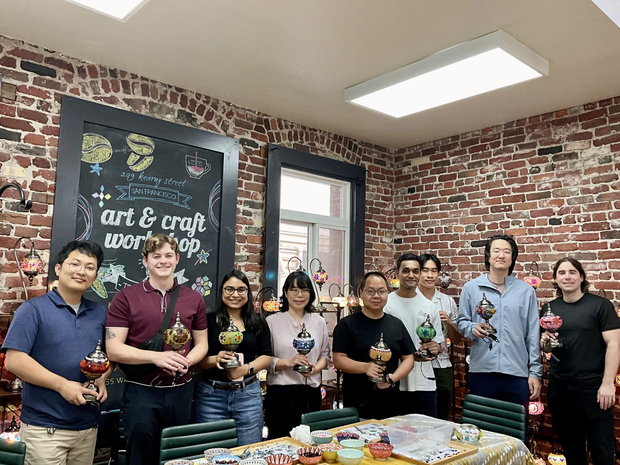 Group of ten people standing in front of a brick wall at an art and craft workshop, holding colorful mosaic glass lanterns, with a black chalkboard sign and display of lanterns in the background.