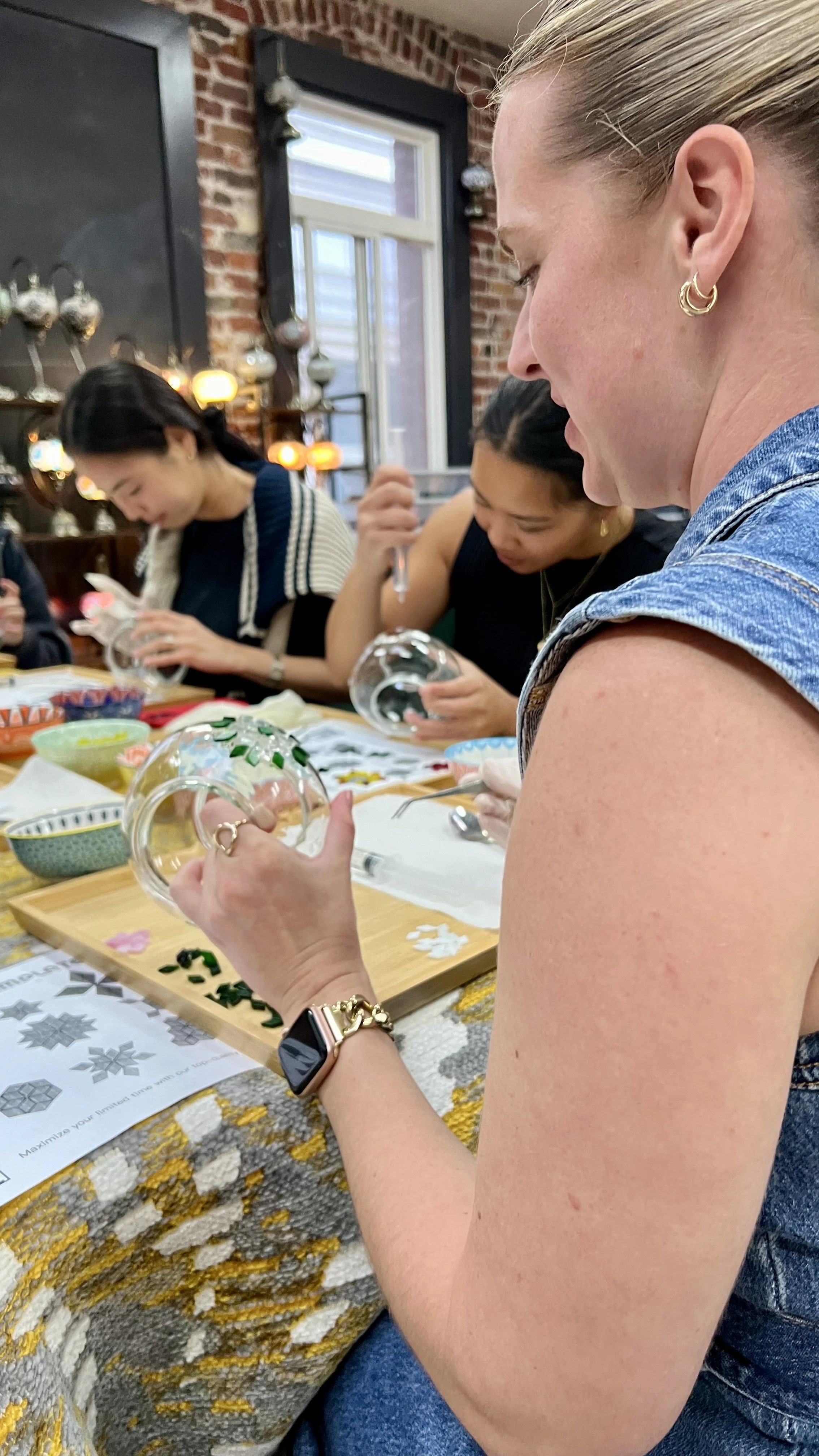 Women gathered around a table decorating glass ornaments, with some focusing on their craft and others looking down at the decorations. The room has a brick wall and a window with natural light.