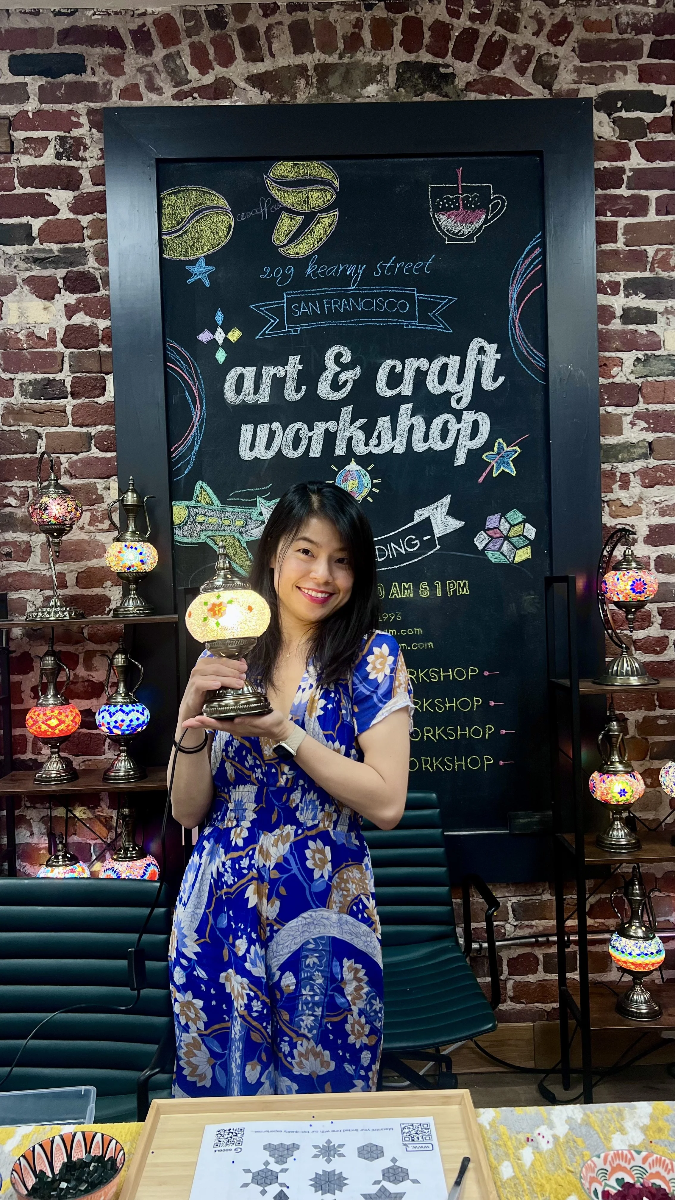 Woman smiling and holding a colorful mosaic glass lamp at an art and craft workshop with a brick wall and decorated lamps in the background.