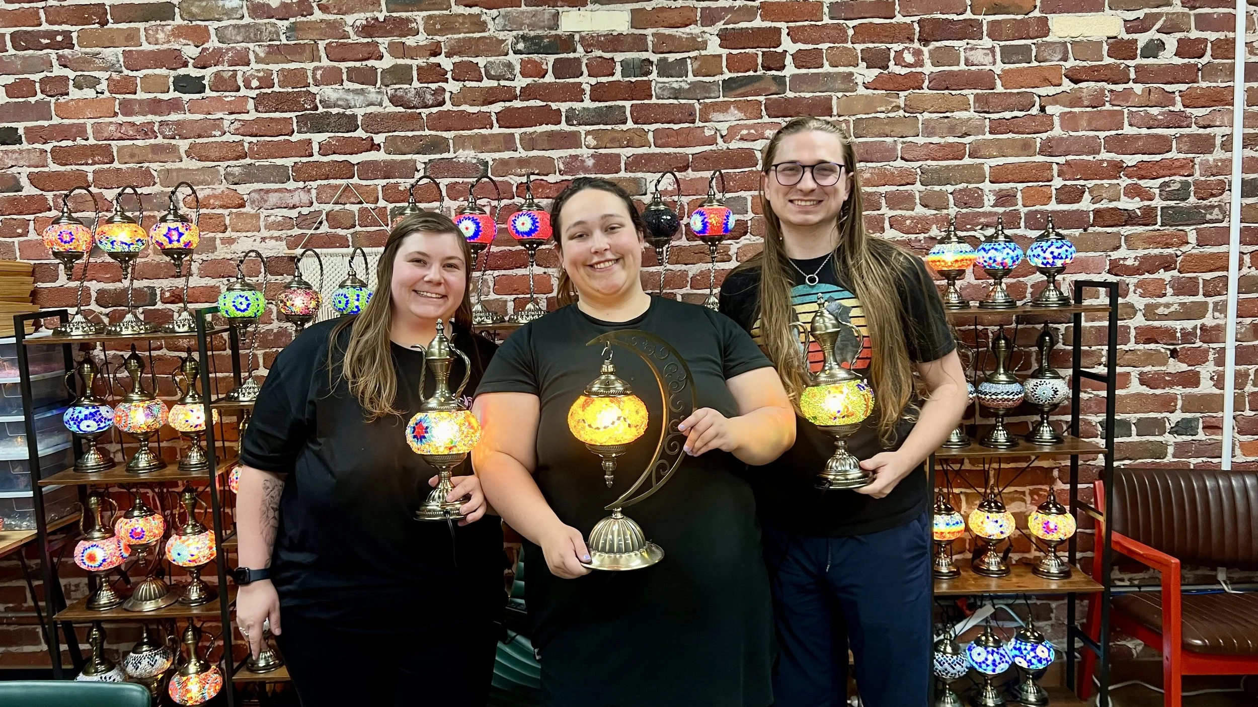 Three women standing in front of a wall with colorful mosaic lamps on shelves, holding lamps and smiling at the camera.