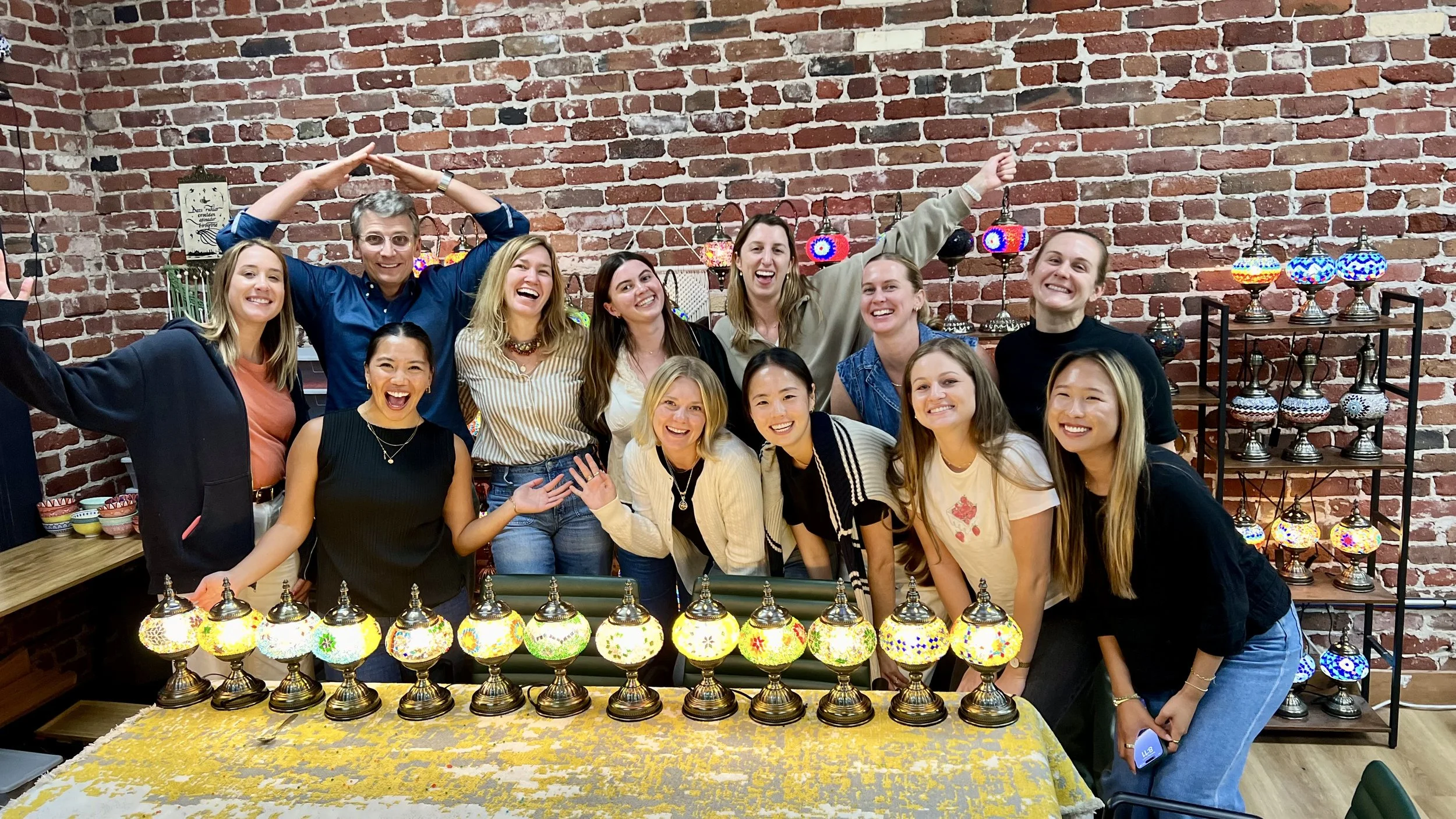 A group of twelve women and one man smiling and posing together indoors in front of a brick wall, surrounded by colorful mosaic lamps and lanterns on tables and shelves.