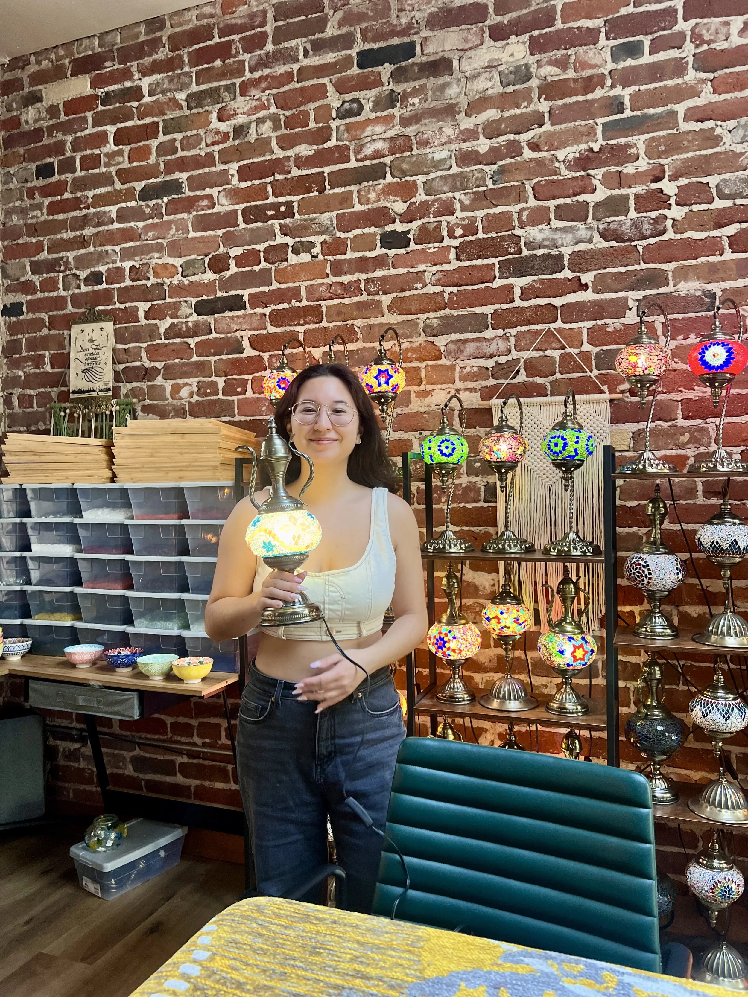 A woman with glasses and wearing a sleeveless beige top and black jeans is holding a colorful mosaic table lamp in a craft shop. Behind her are shelves displaying various mosaic lamps and a wall with a brick pattern.
