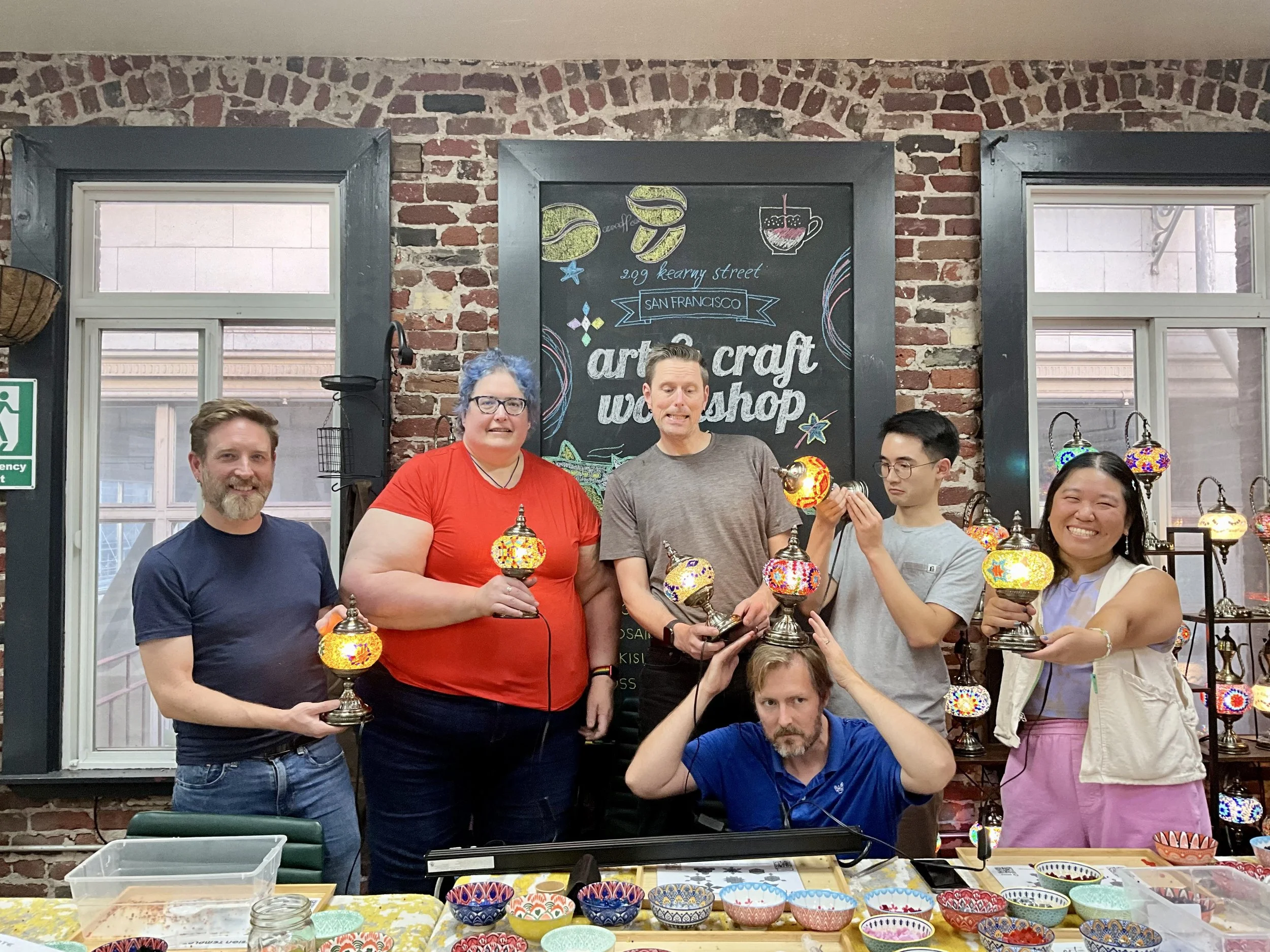 Group of six people standing behind a table with colorful mosaic lamps, holding and displaying various lamps in a craft workshop setting.