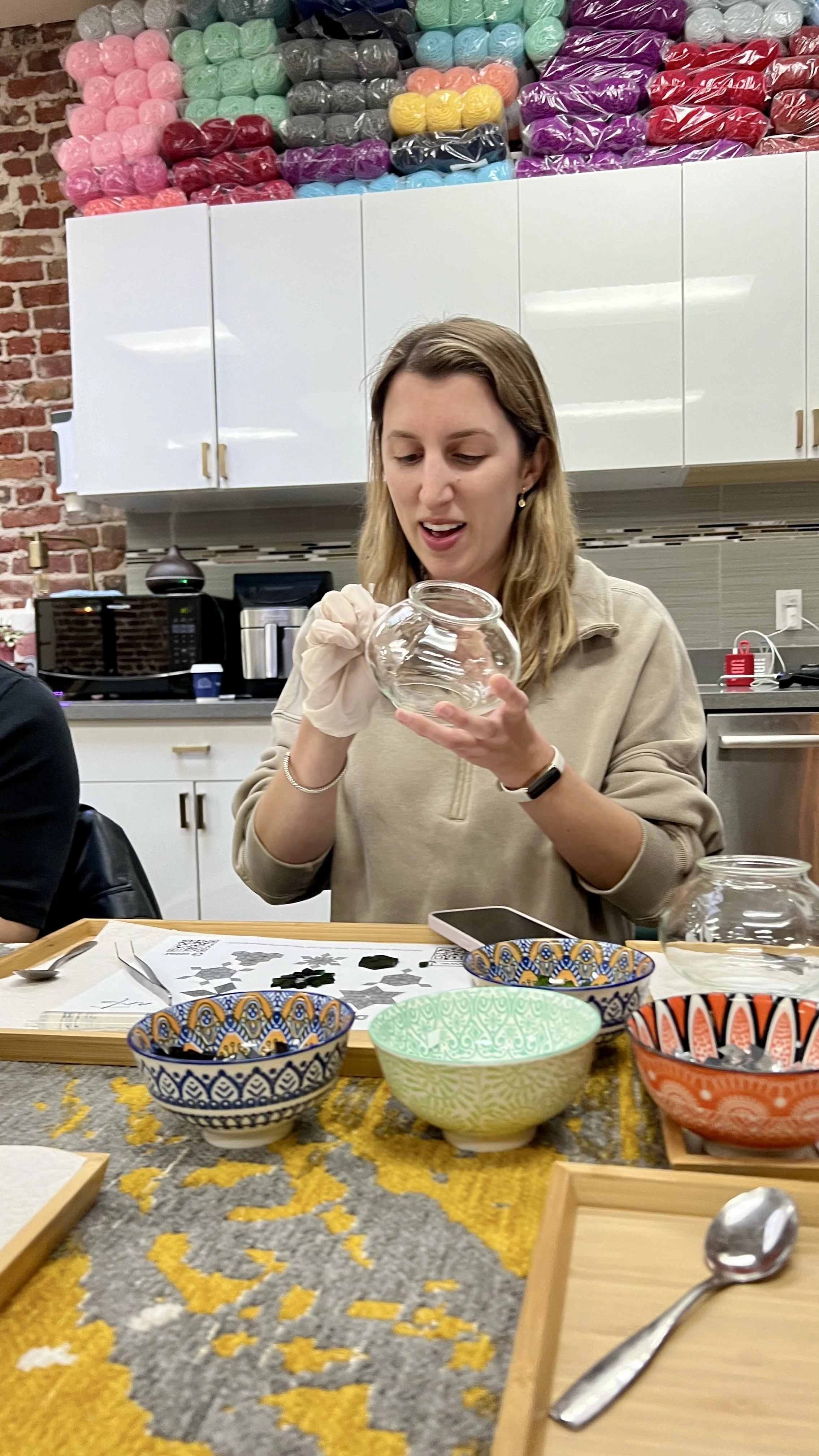 A woman sitting at a table with colorful bowls and a glass jar, holding a small round glass jar while looking at it. The background shows a Kitchen with white cabinets, a brick wall, and shelves filled with yarn balls in various colors.