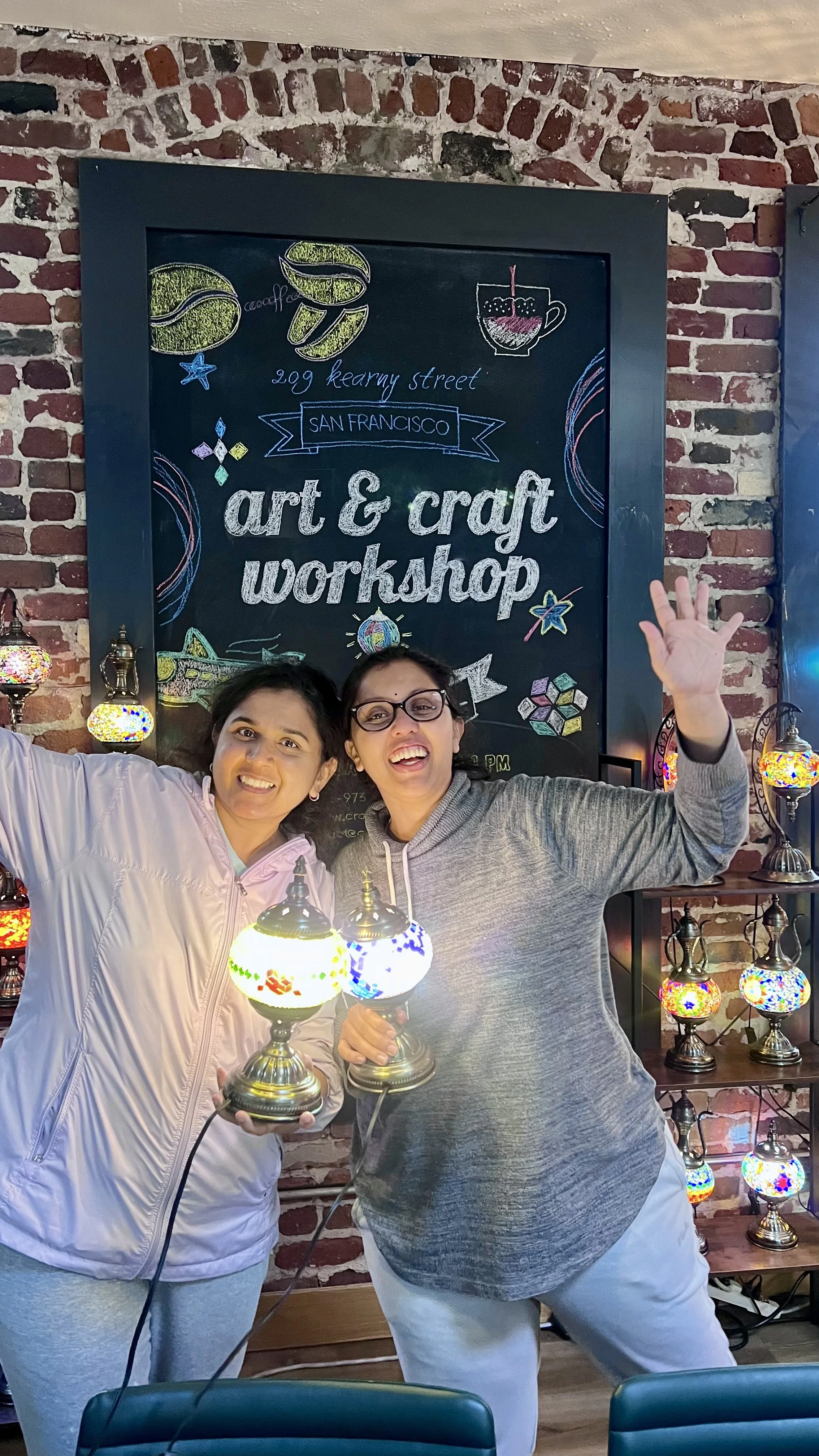 Two women smiling and holding colorful lamps at an art and craft workshop in front of a chalkboard with drawings of coffee cups, cookies, and art supplies.