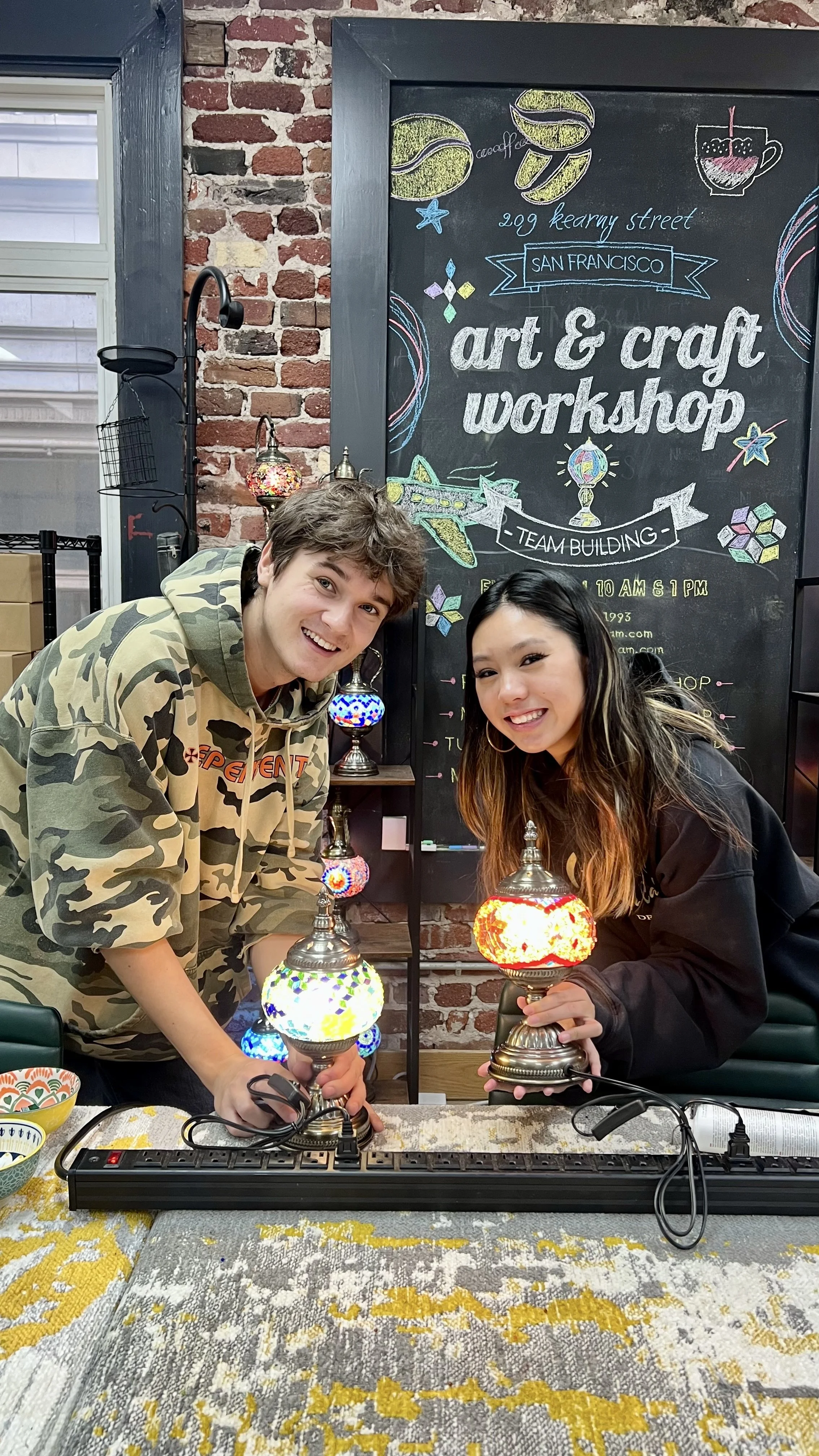 Two smiling people holding colorful mosaic table lamps at a craft workshop in a room with a chalkboard sign that reads 'art & craft workshop' and a brick wall in the background.