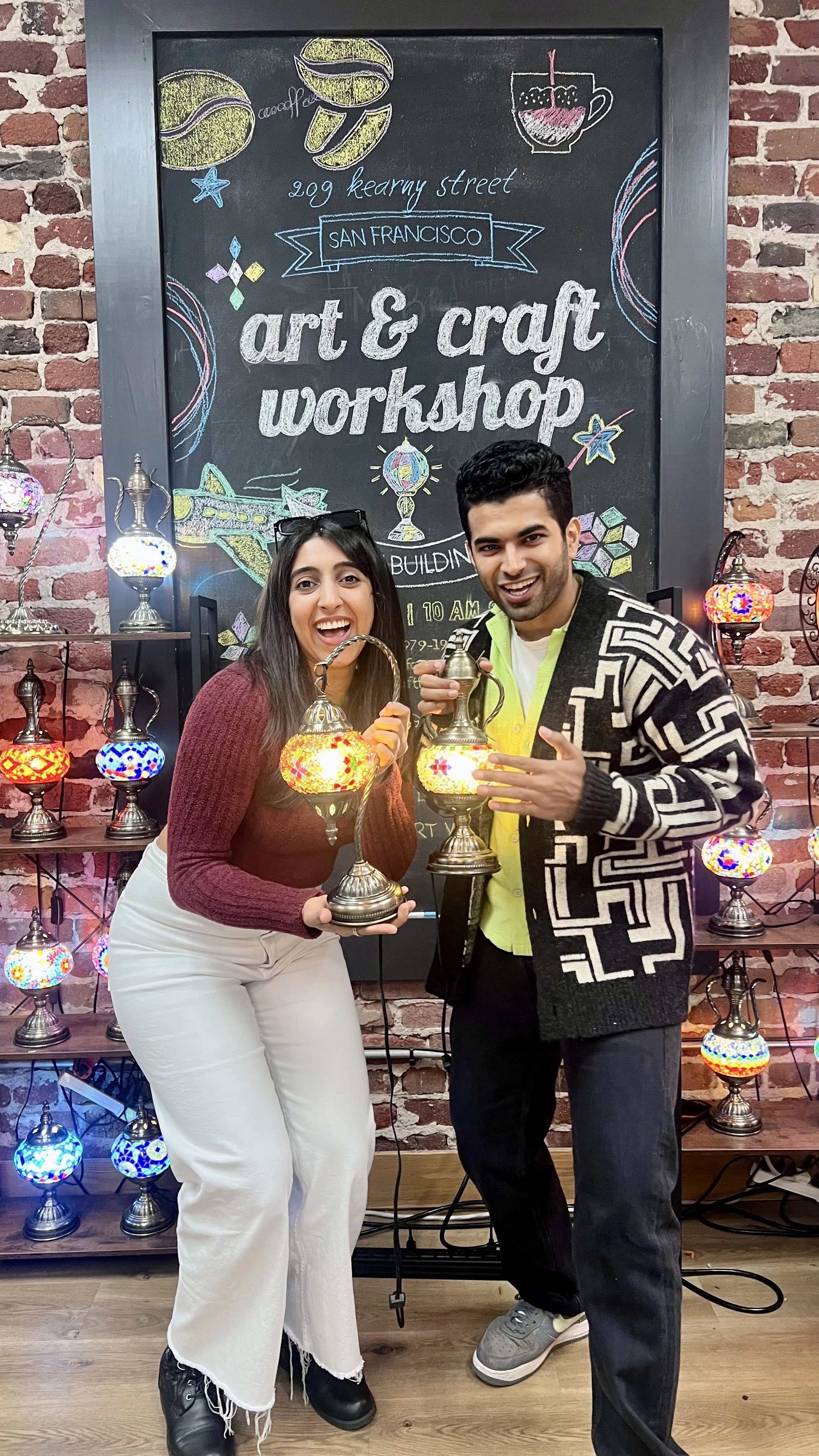 Two people smiling and holding decorative lamps at an art and craft workshop in front of a chalkboard sign with colorful drawings.