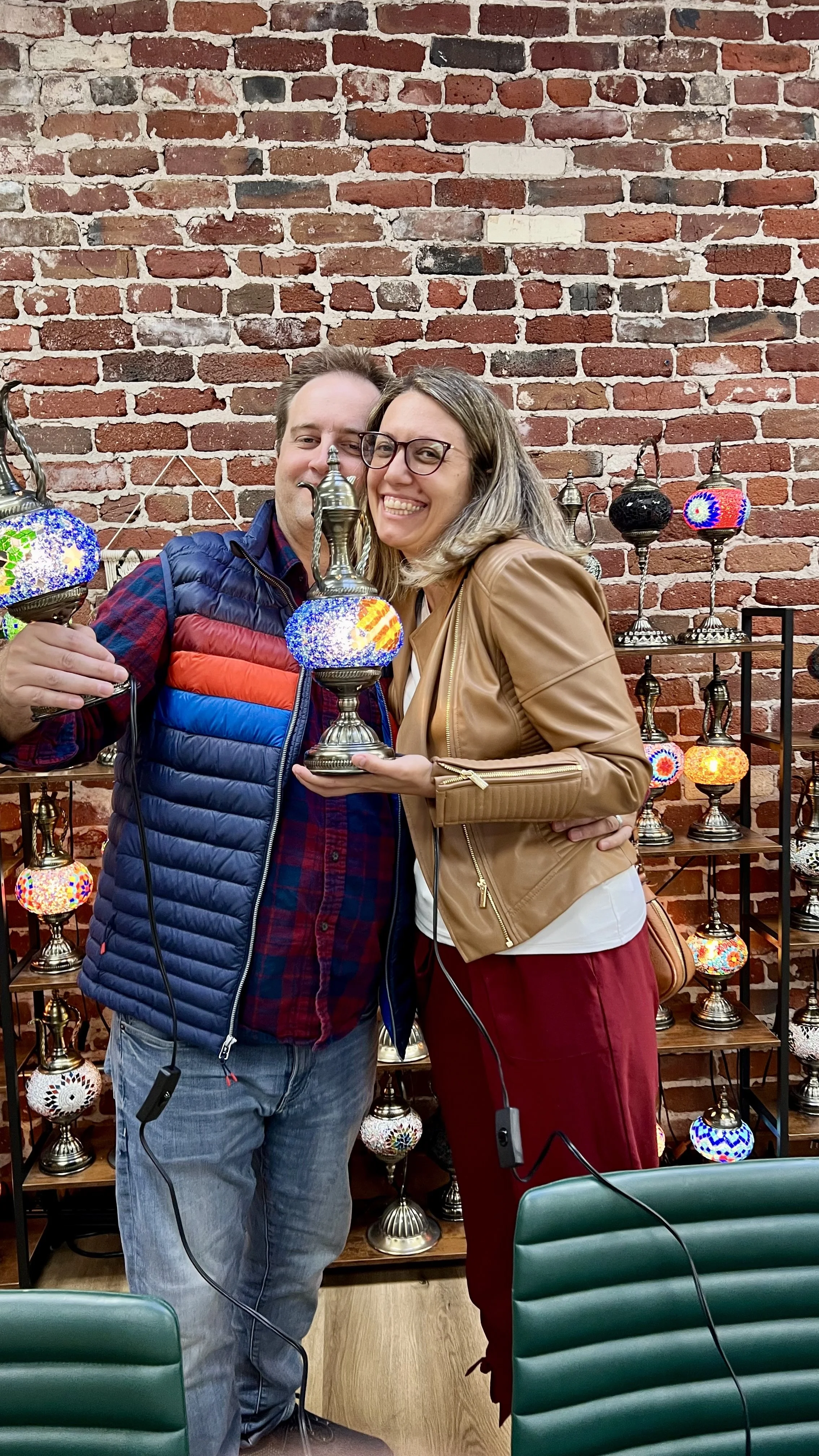 A man and woman smiling and holding colorful Turkish-style lamps in front of a brick wall with display shelves of similar lamps.