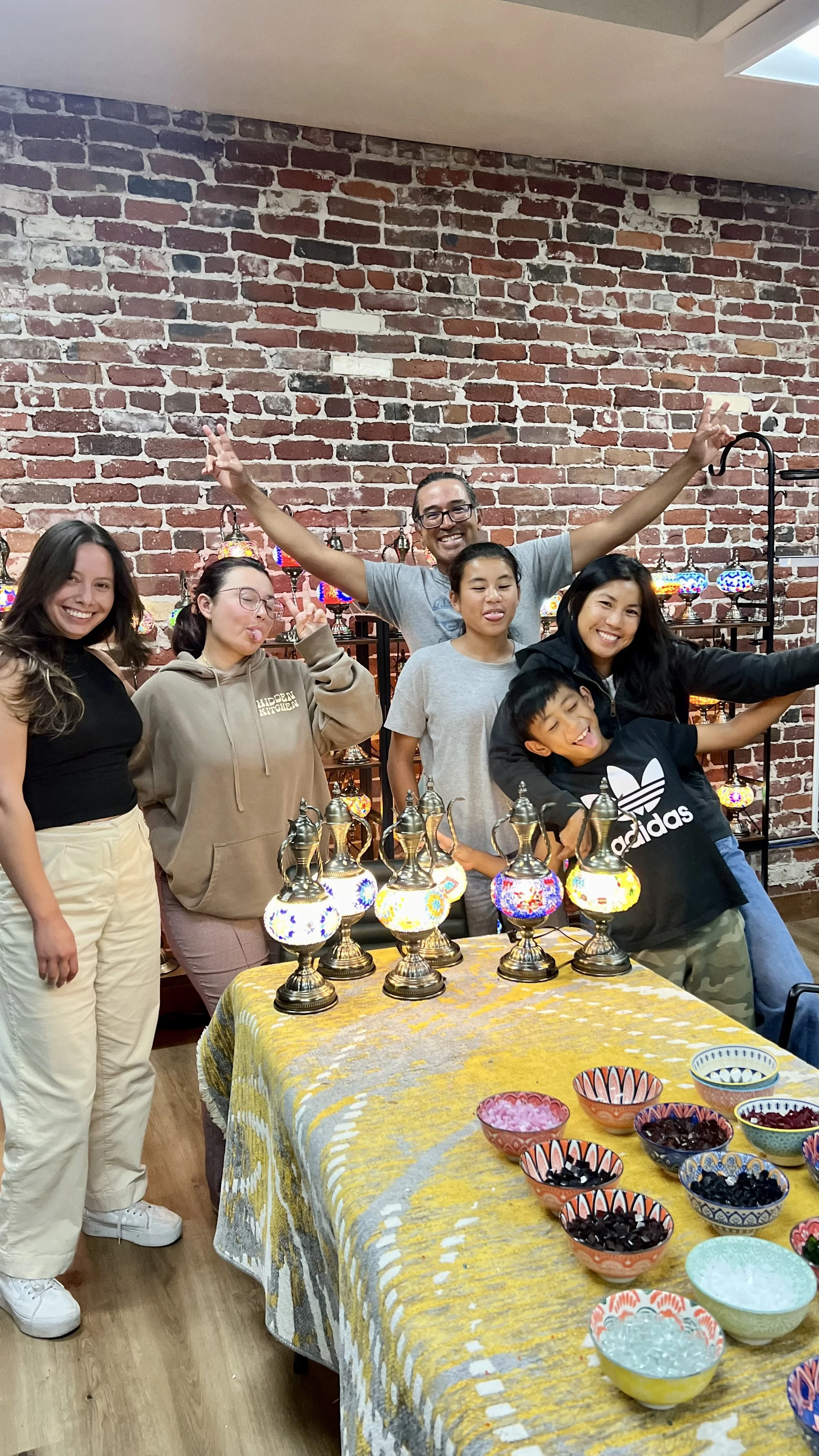 Group of six people posing behind a table with colorful lamps and bowls, with a brick wall in the background.