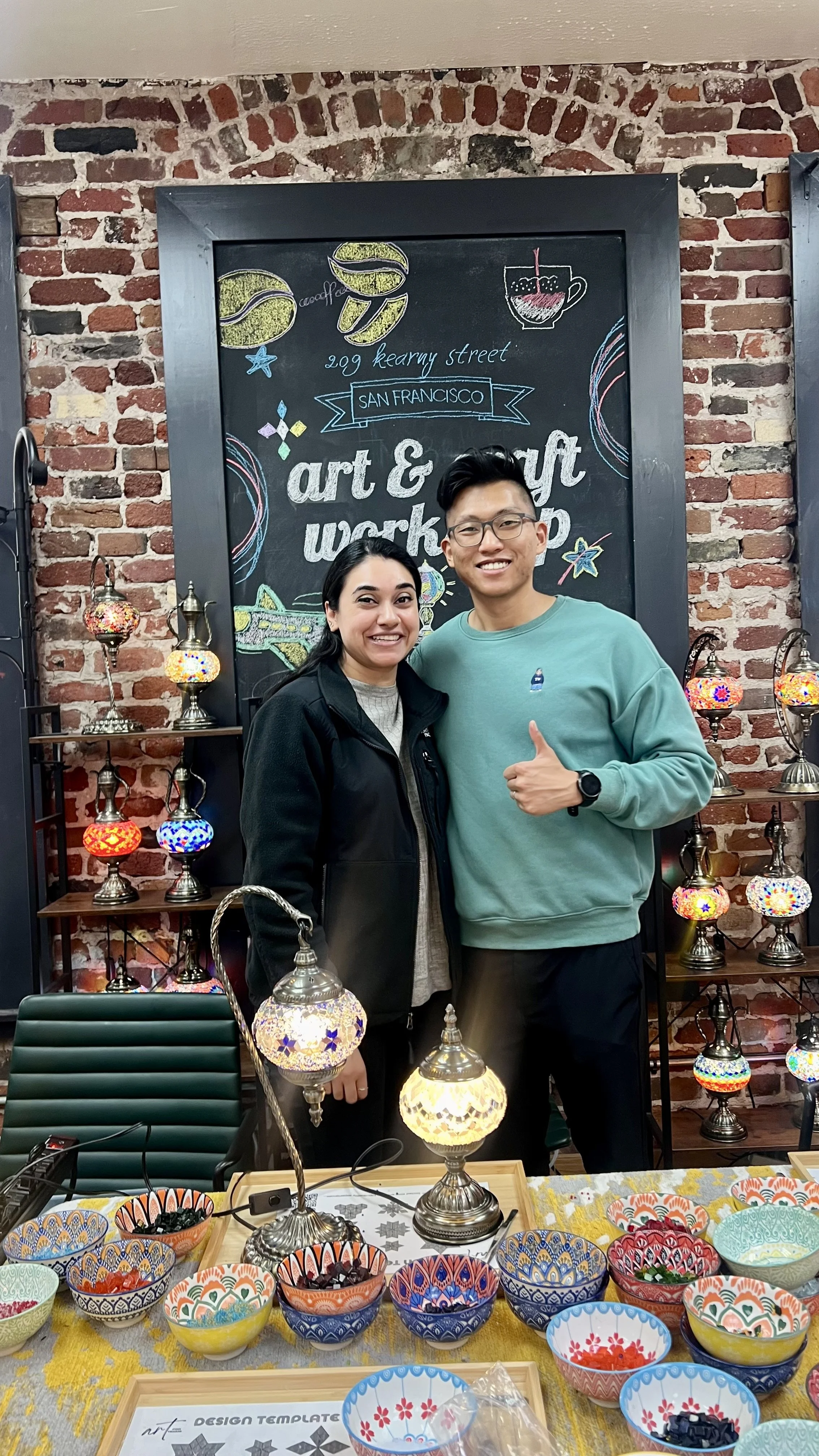 Two people smiling and standing together in front of a blackboard at an art and craft workshop, with colorful mosaic lamps and bowls on the table in front of them and a brick wall behind.