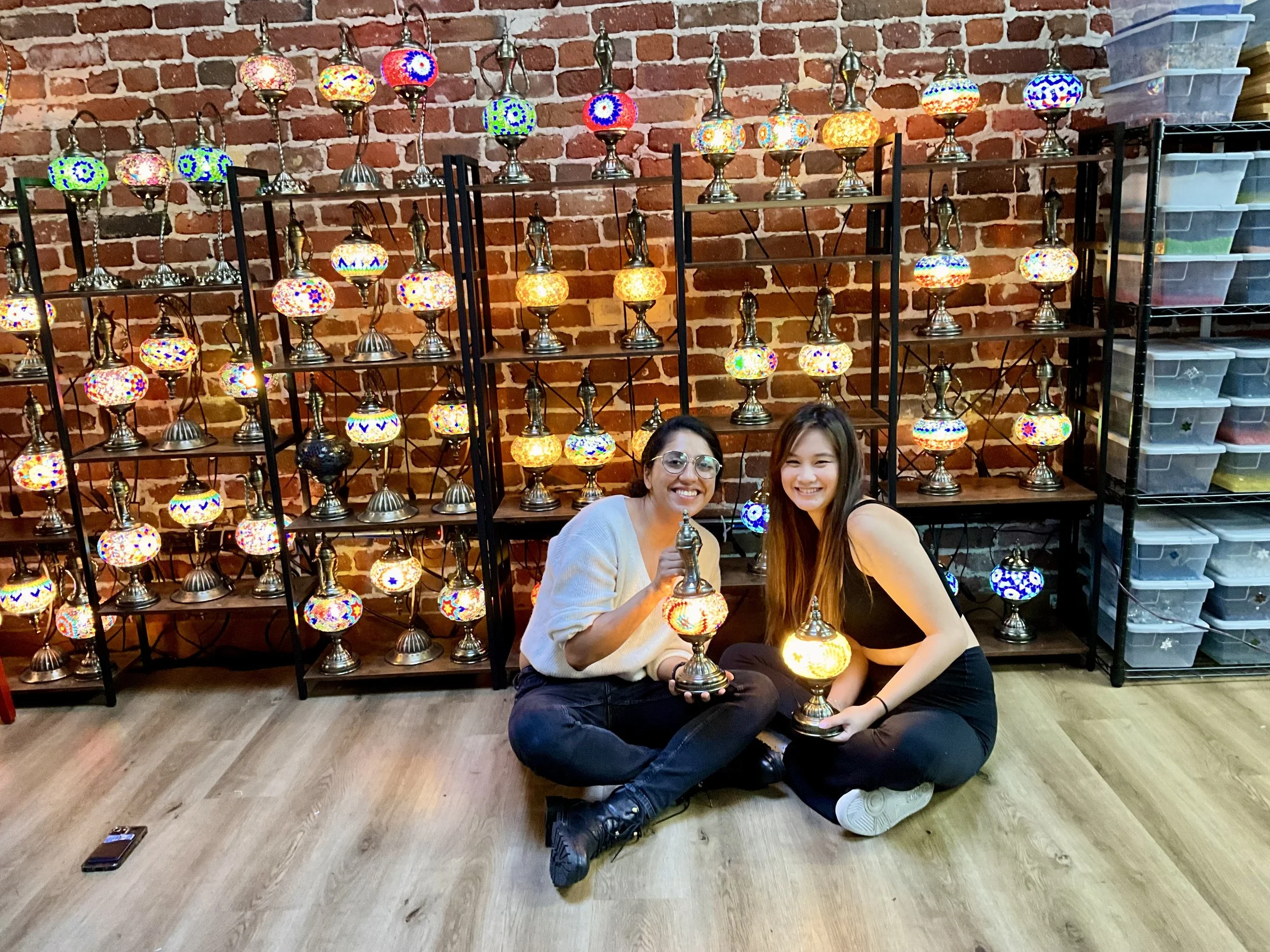 Two women sitting on the floor in front of display shelves filled with colorful mosaic lamps, with exposed brick wall in the background.