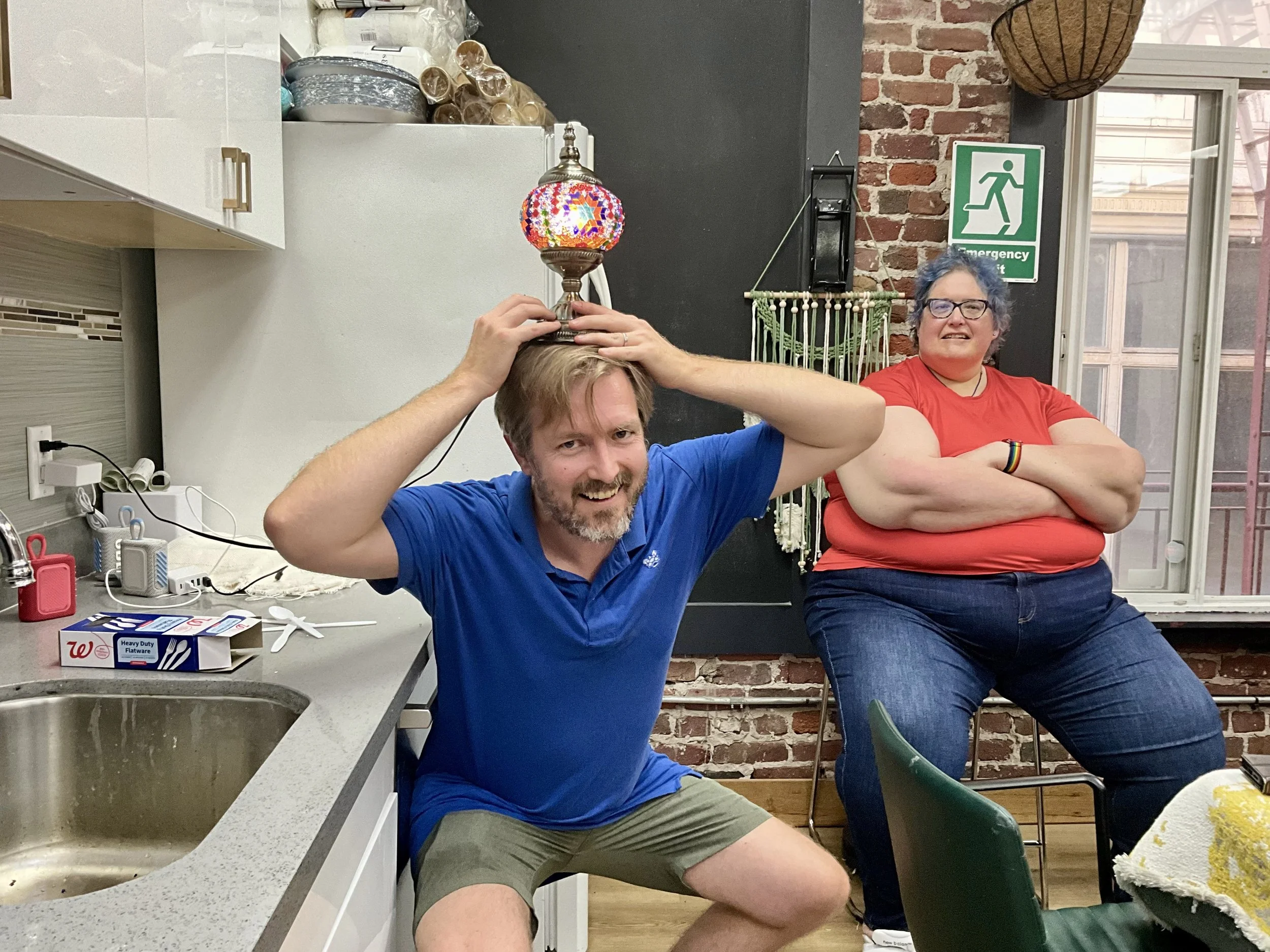 A man in a blue shirt and shorts balancing a colorful lamp on his head, with a woman in a red shirt sitting behind him with her arms crossed, smiling in a kitchen with brick walls and a green emergency exit sign.