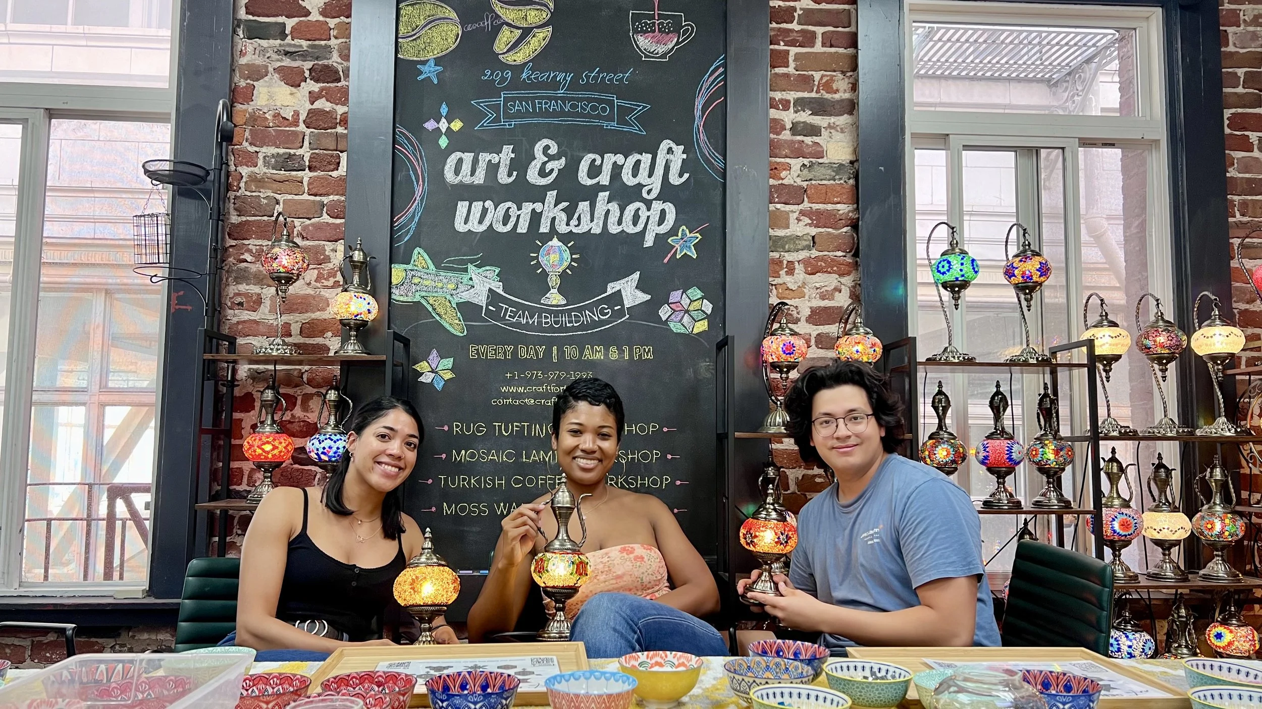 Three smiling people seated at a table in an art and craft workshop, holding colorful mosaic lamps. Behind them is a chalkboard sign with details of the workshop, and shelves displaying various mosaic lamps against a brick wall.