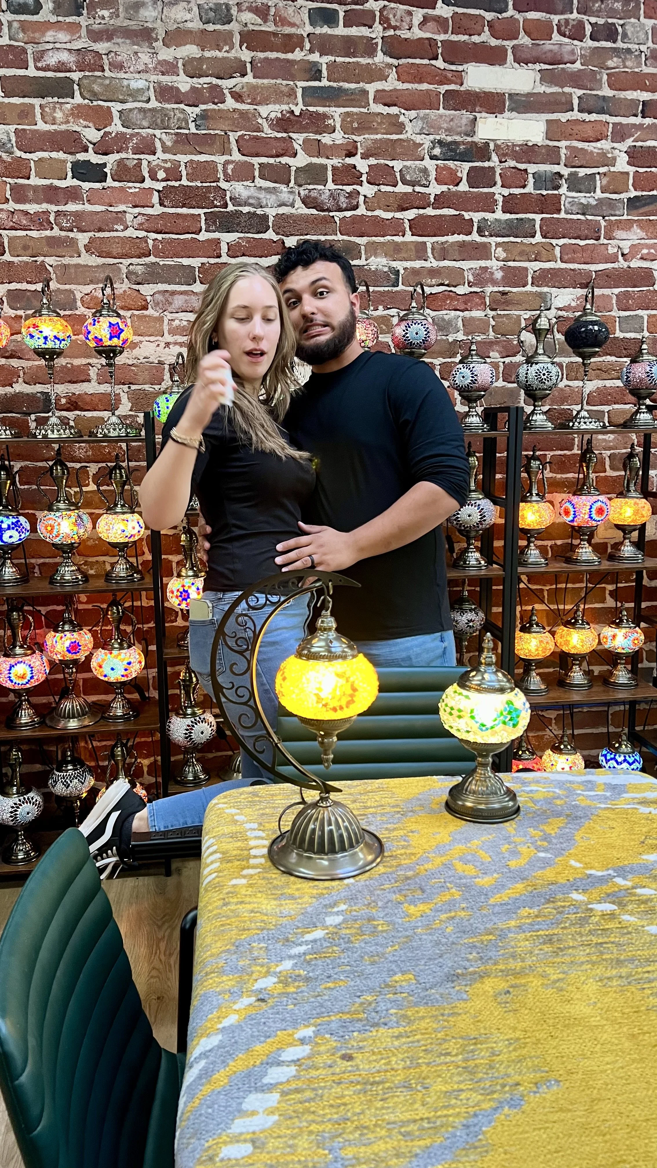 A man and woman posing in front of a display of colorful Turkish mosaic lamps, with a brick wall background and a yellow and grey patterned table in the foreground.
