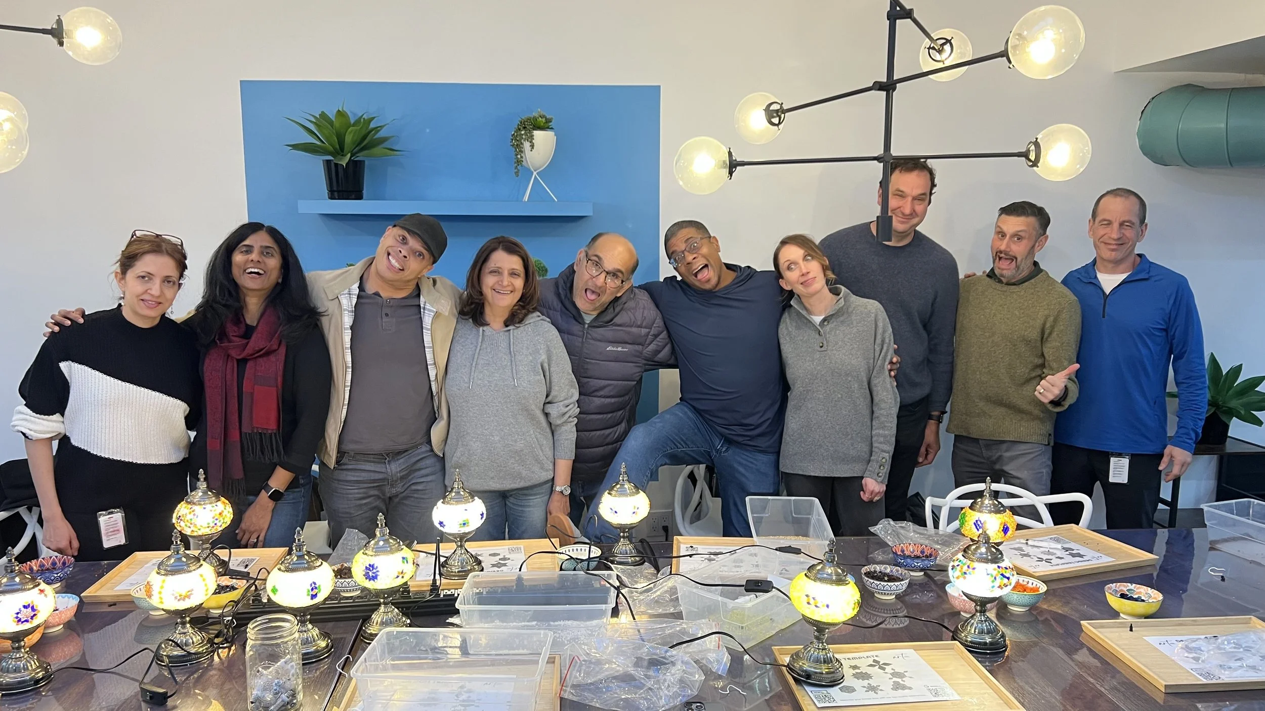 Group of ten smiling people standing together behind a table with colorful mosaic lamps and bowls, in a room with modern lighting and blue wall accents.