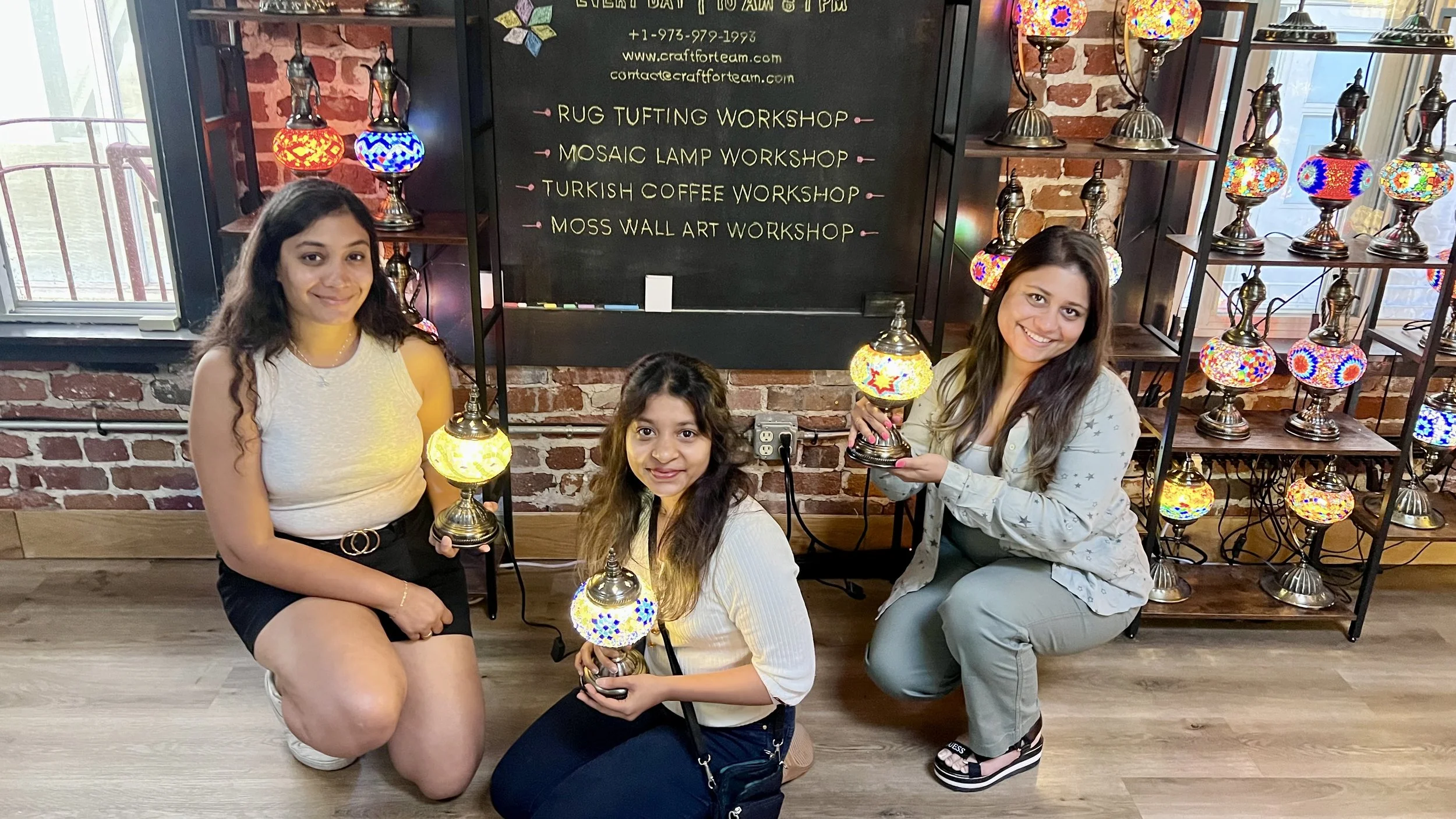 Three women kneeling on the floor in front of a brick wall with a blackboard, holding colorful mosaic lamps, with more mosaic lamps displayed on shelves behind them, inside a craft workshop.