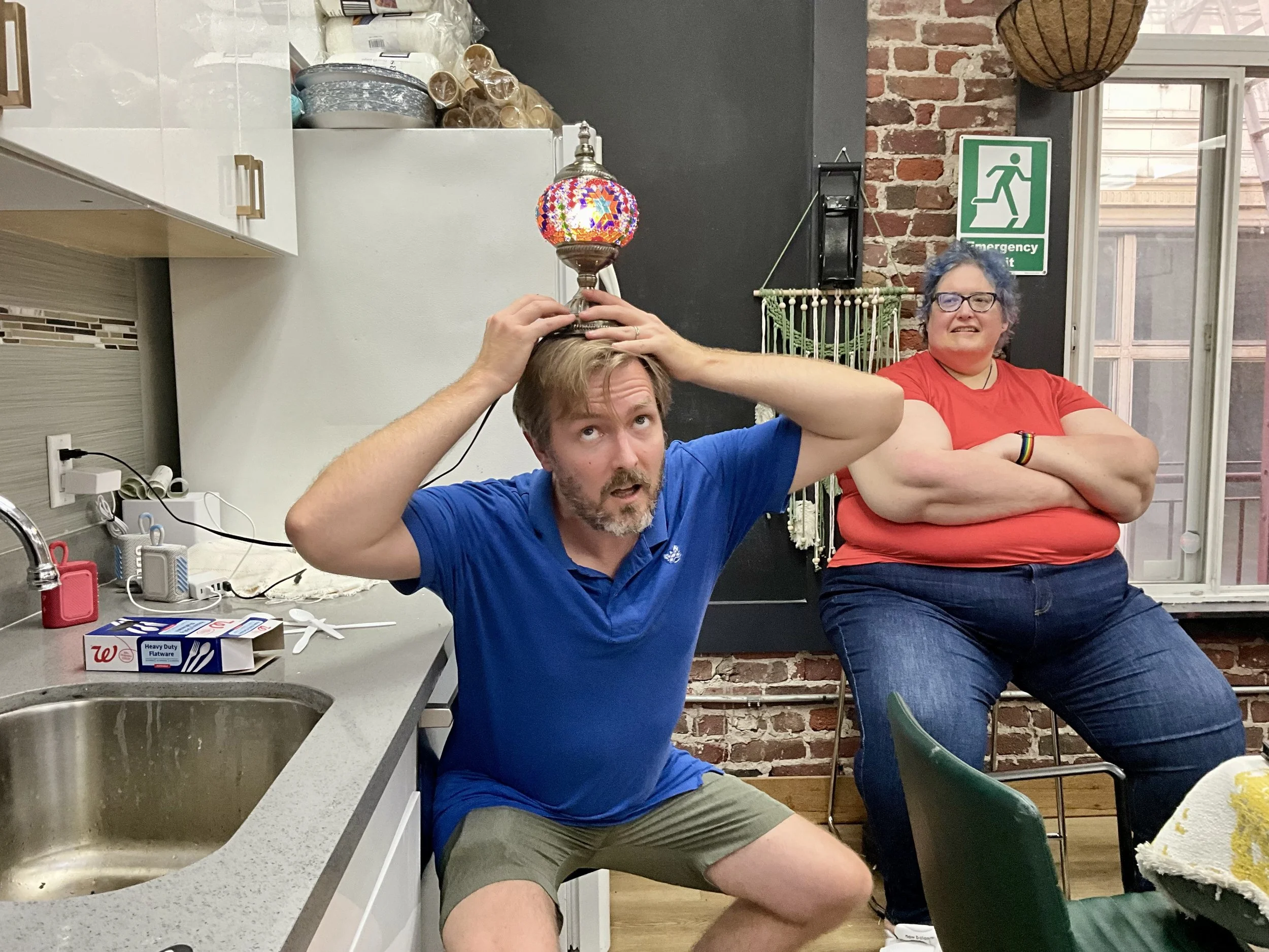 A man sitting in a kitchen with a lampshade on his head, holding it with both hands, while a woman in the background with her arms crossed and smiling.