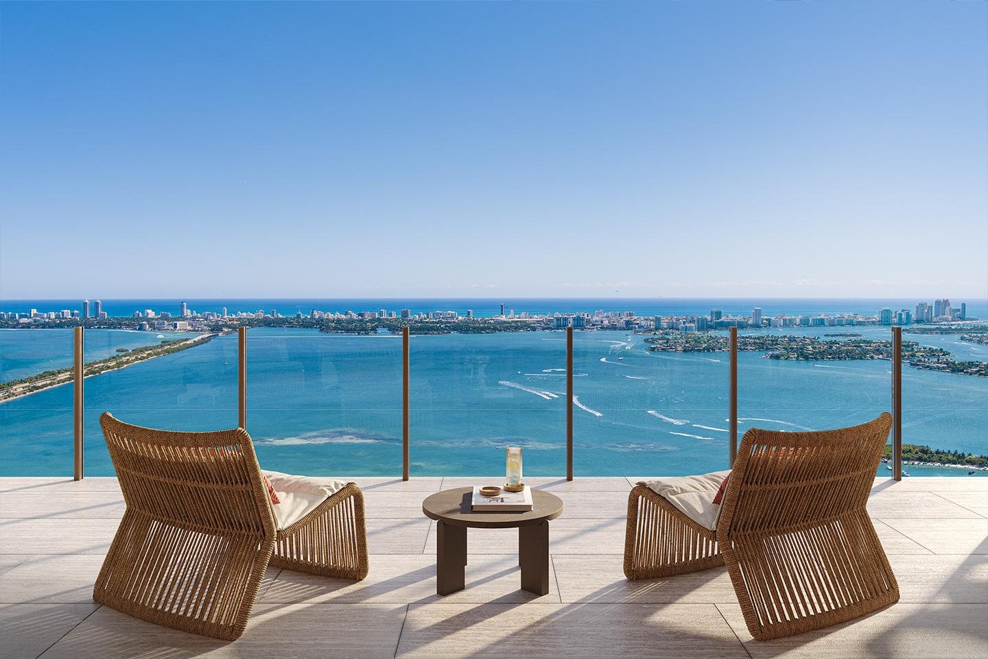 Balcony with two wicker chairs and a small table, overlooking a large body of water and a city skyline in the distance under a clear blue sky.