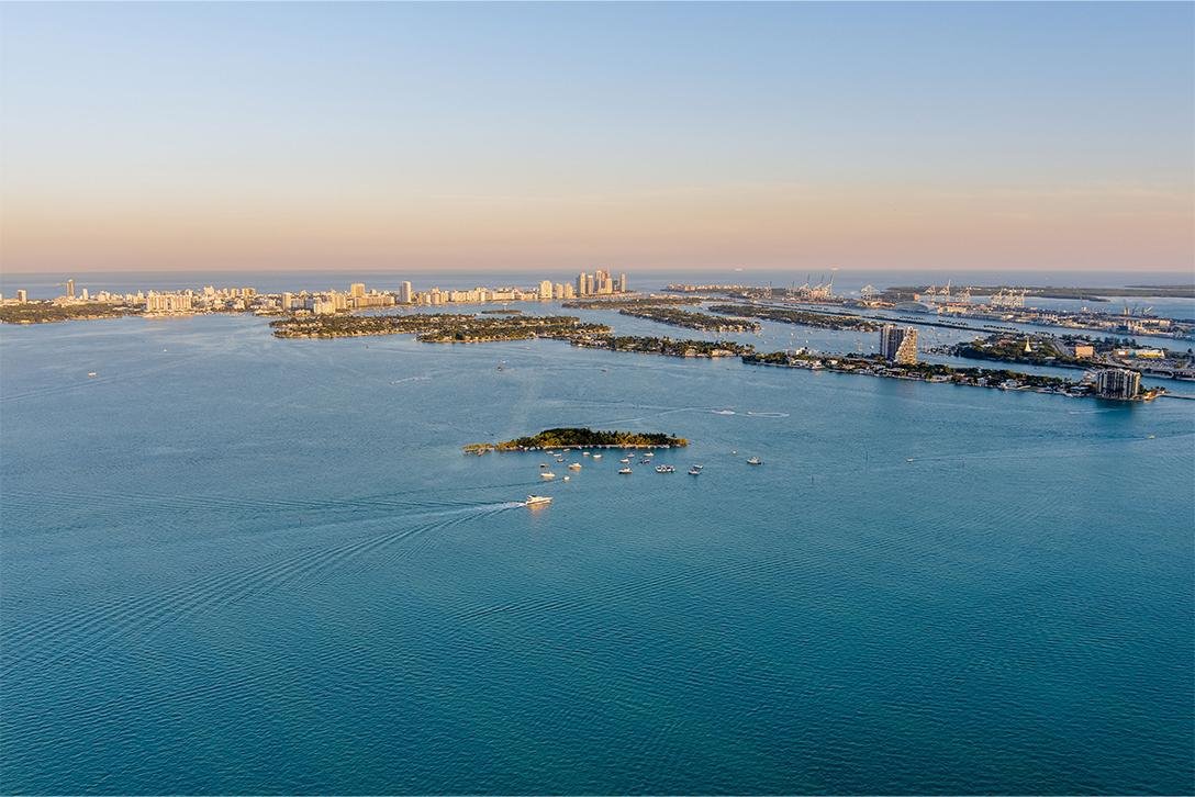 Aerial view of a large body of water with a small island and boats, with a city skyline and harbor in the background.