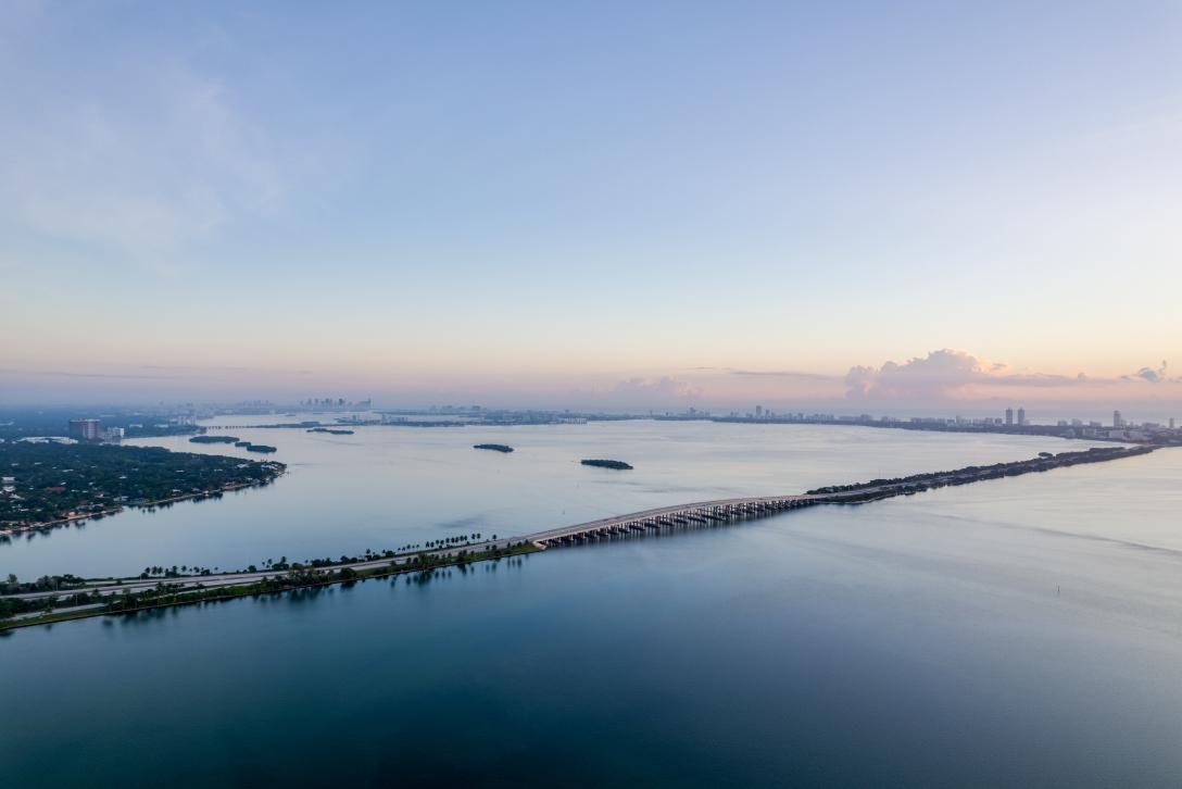 Aerial view of a cityscape with a large body of water, a long bridge connecting landmasses, and high-rise buildings in the distance.