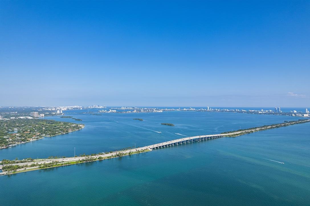Aerial view of a large body of water with a bridge crossing over it, city skyline in the distance, and lush green landscape on one side.