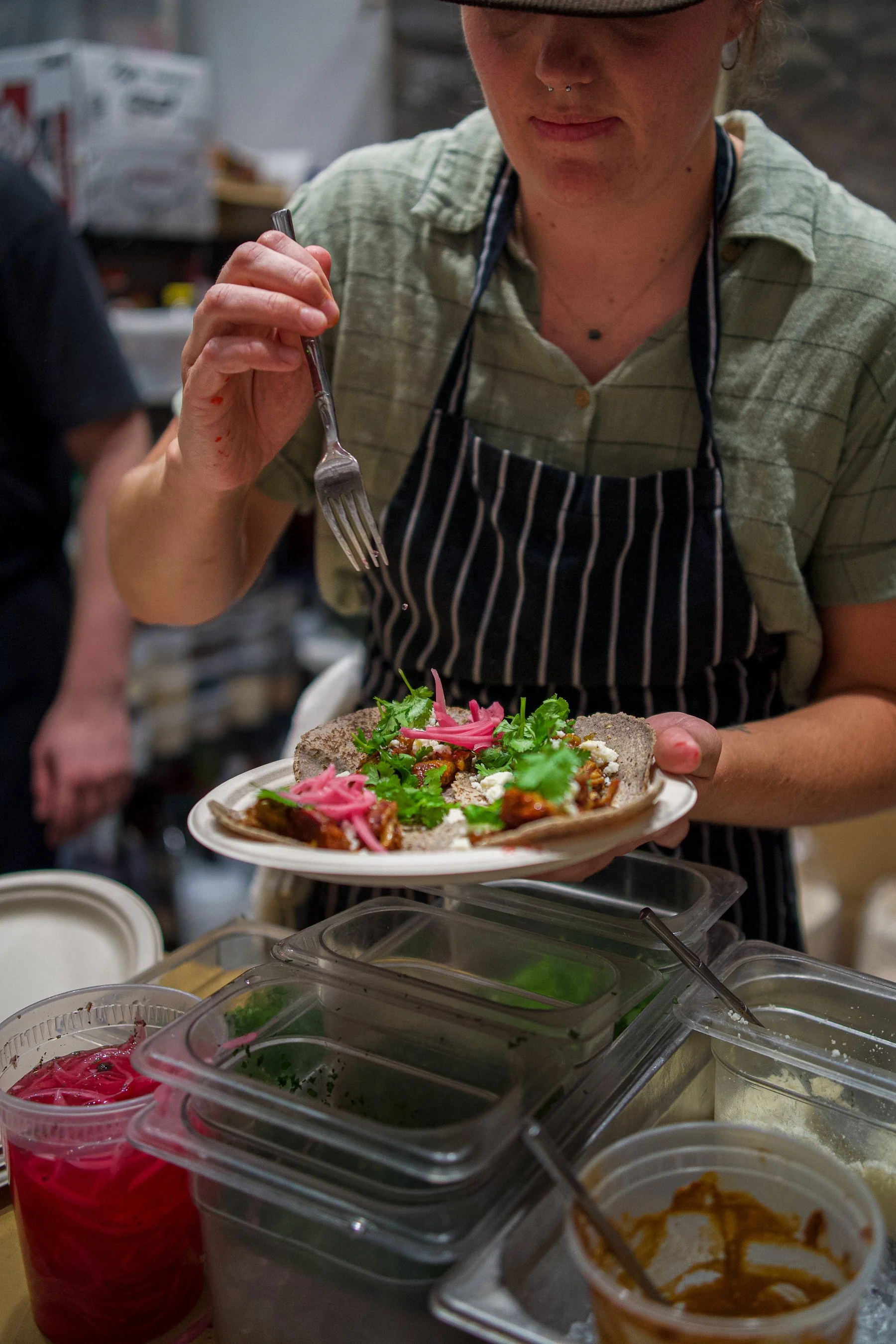 A woman in a striped apron holding a plate of tacos, topped with cilantro, pickled onions, and sauce, in a kitchen or food preparation area surrounded by various containers of ingredients.