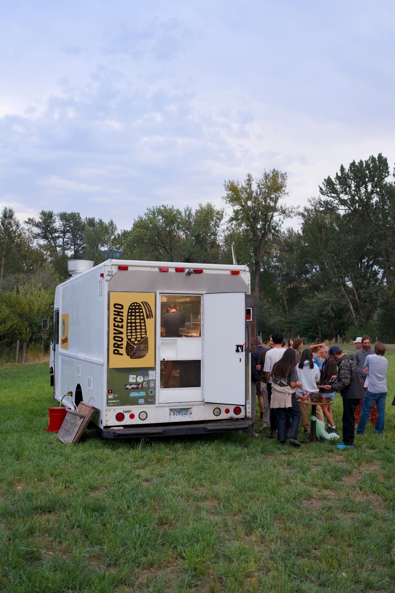 A mobile food truck serving tacos by Provecho, with a group of people gathered around outdoors in a grassy area. The truck has a yellow logo with a rustic corn print.