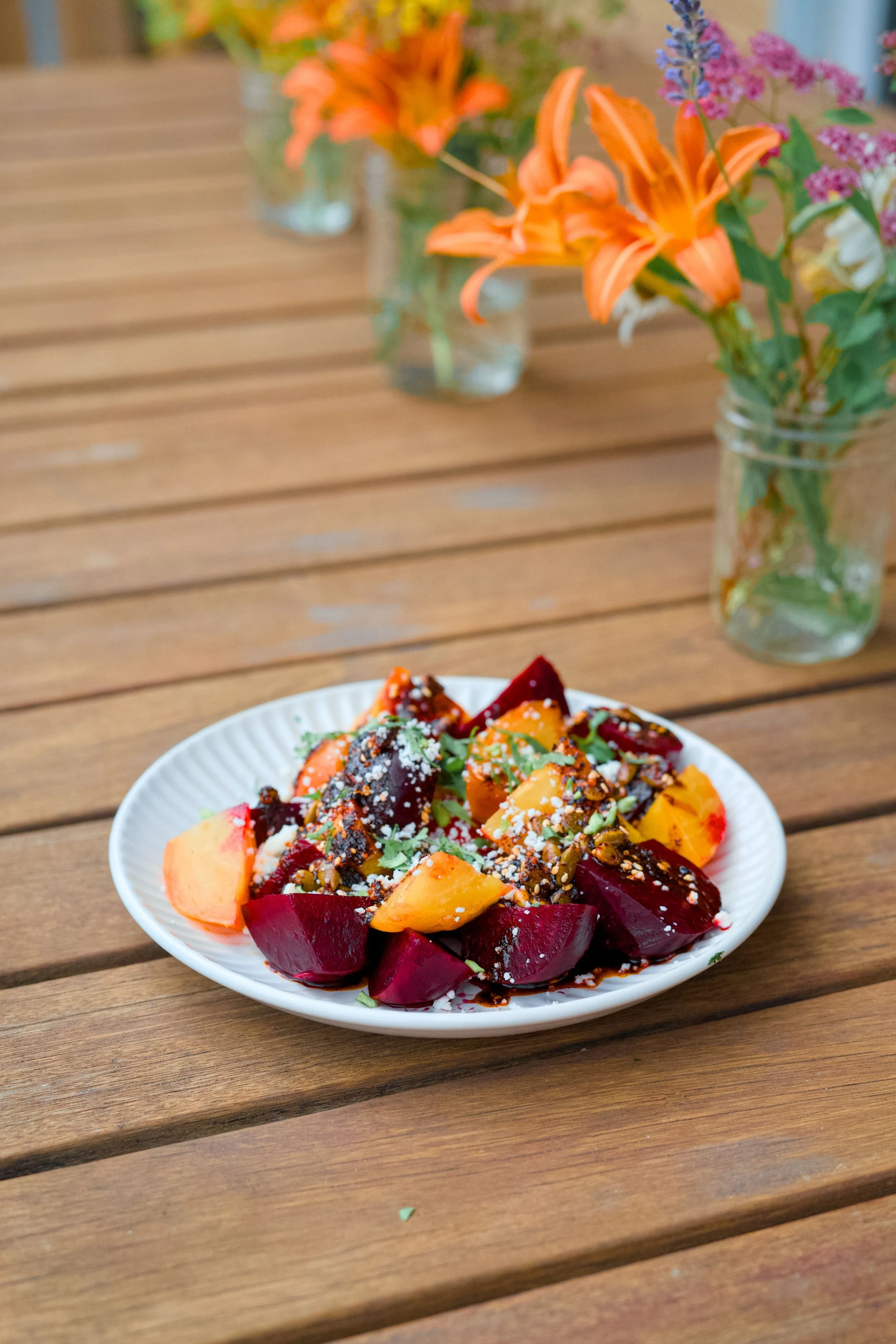 Vegetable salad with red and gold beets and herbs on a white plate, with orange lilies and purple flowers in glass jars in the background on a wooden table.