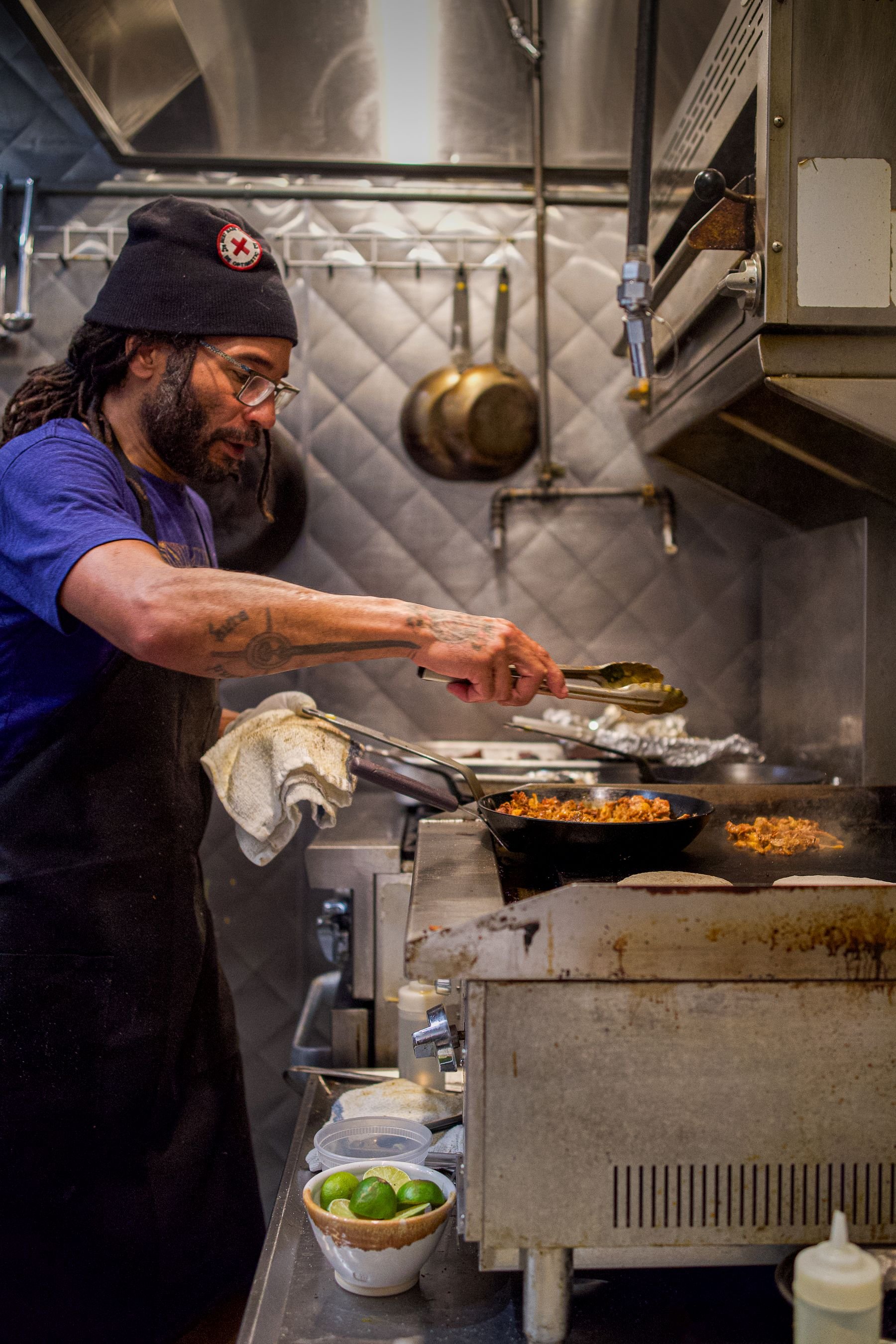 A man cooking at a stove in a commercial kitchen, using tongs to cook food in a skillet. There are limes in a bowl and various kitchen tools around him.