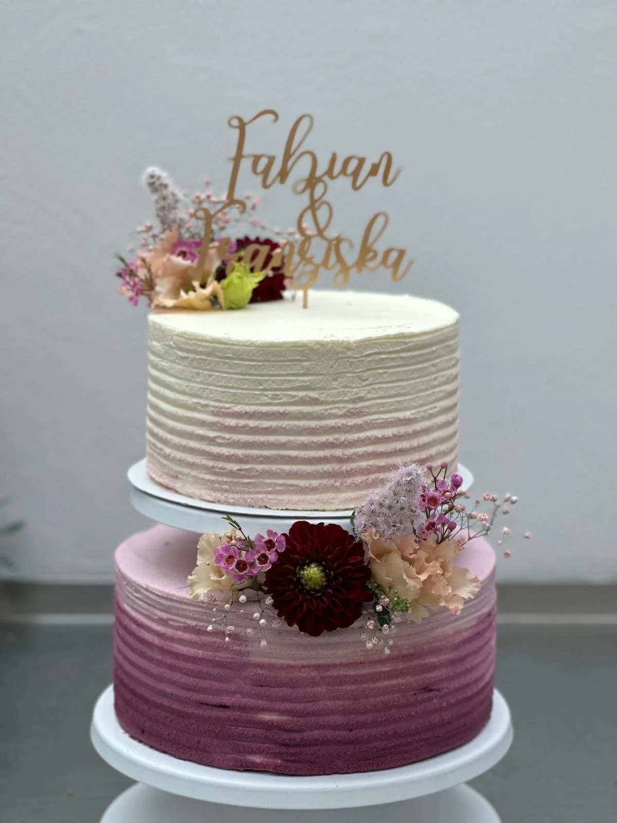 Two-tiered wedding cake with pink and white ombre frosting, decorated with pink, red, and cream flowers, and a gold topper reading 'Fahan & Janiska'.