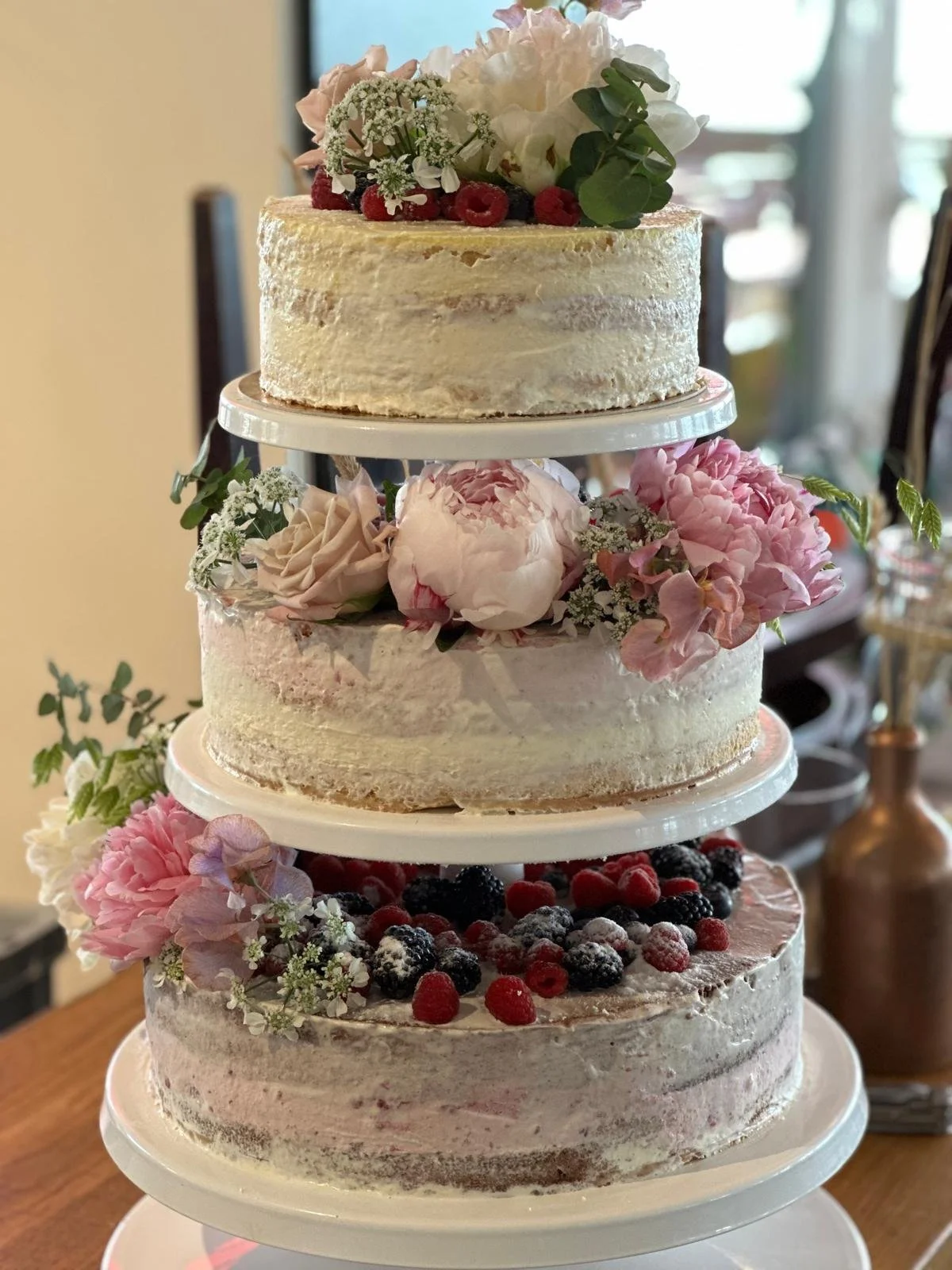 A four-tiered naked cake decorated with pink and white flowers, and topped with berries. The cake has a rustic style with minimal frosting, showcasing layers of sponge and cream.