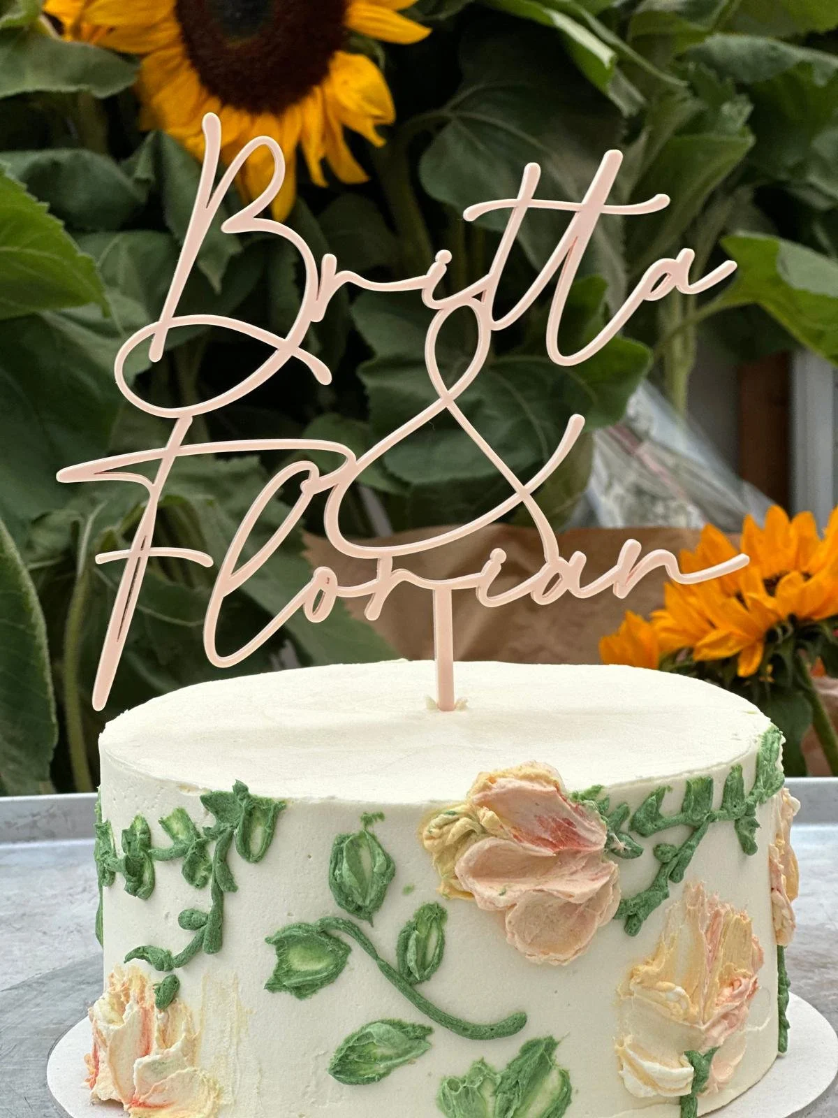 White birthday cake with floral decorations and a pink wire topper that reads 'Brittany'. In the background, there are sunflowers and green leaves.