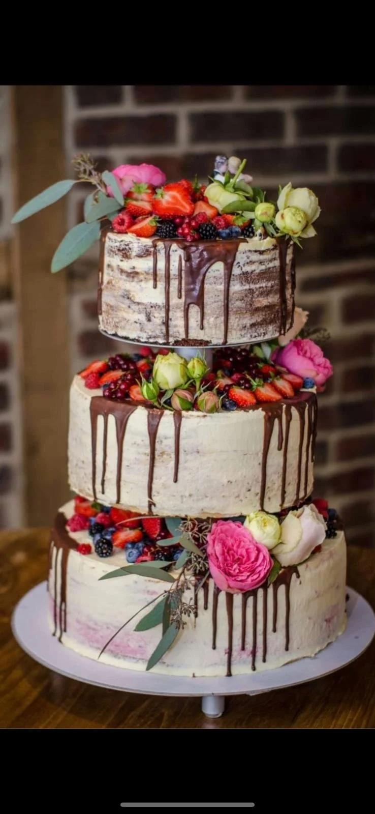 Three-tiered wedding or celebration cake decorated with fresh berries, strawberries, blackberries, raspberries, and pink and white flowers, with chocolate drizzle on each layer, set against a brick wall background.