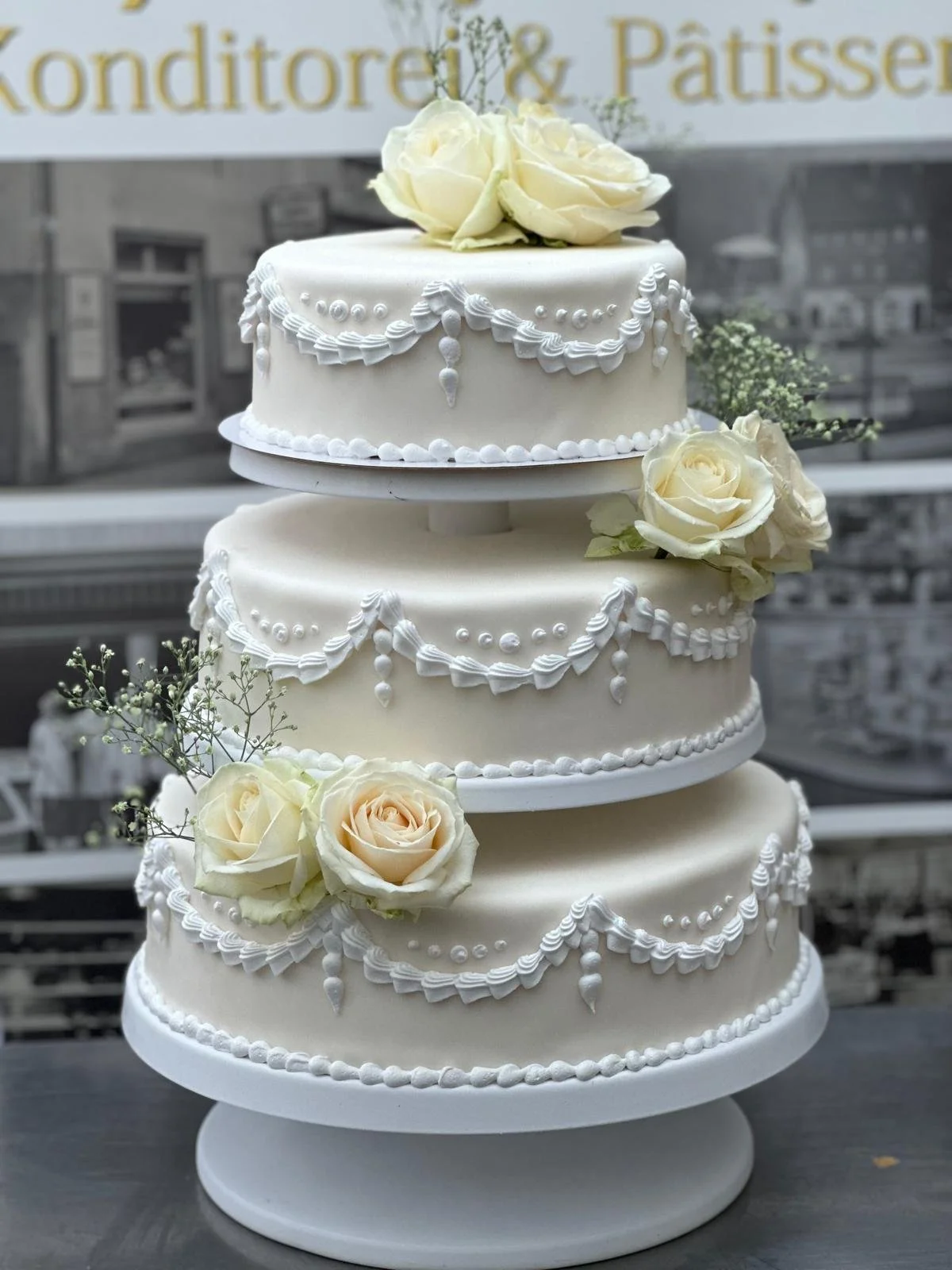Three-tier white wedding cake decorated with white roses and icing garland designs, set on a stand with a blurred background featuring a sign that reads 'Konditorei & Patisserie'.