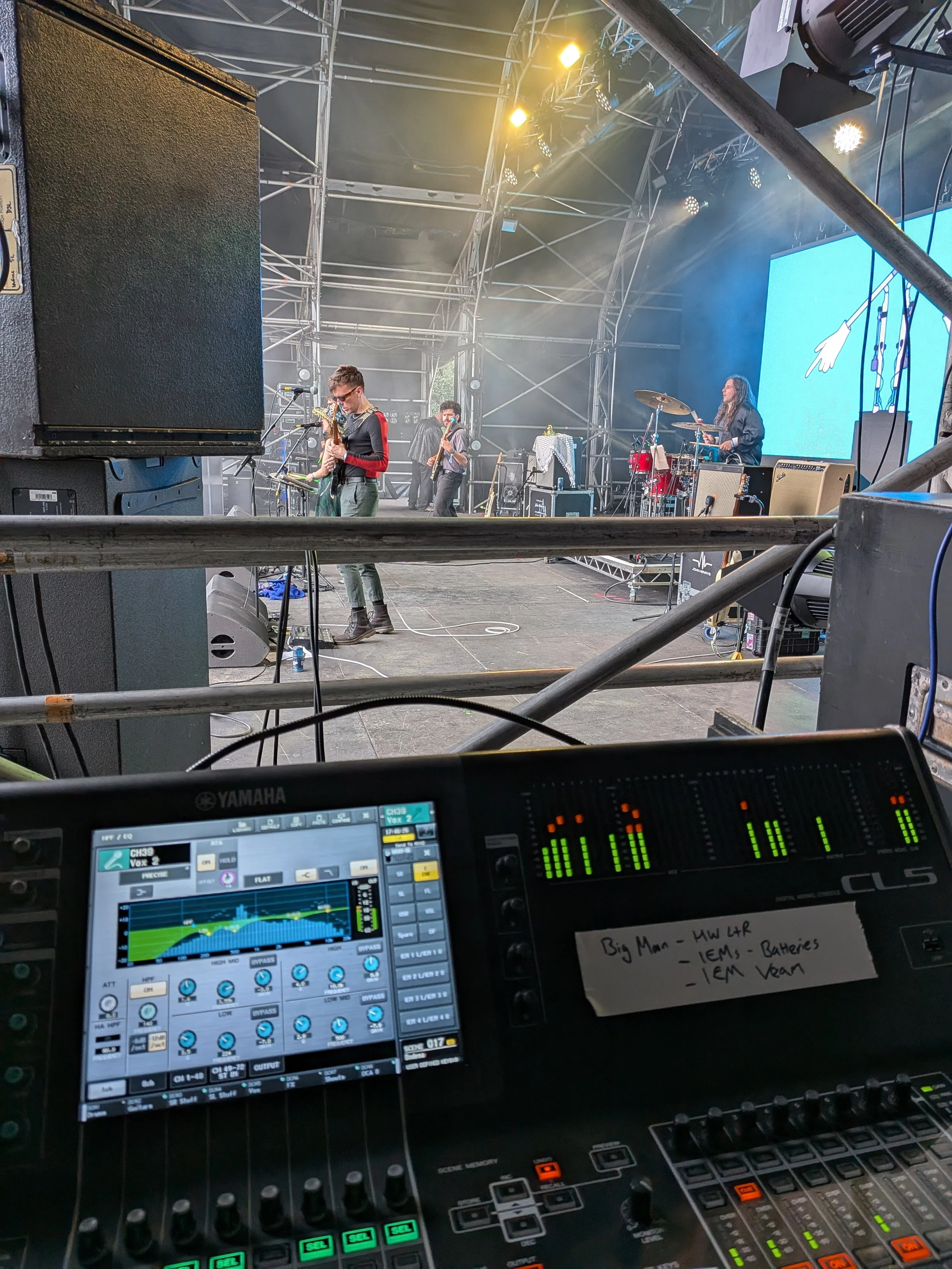 A live music band performing on stage with a guitarist, bassist, and drummer, with a sound mixing board in the foreground.