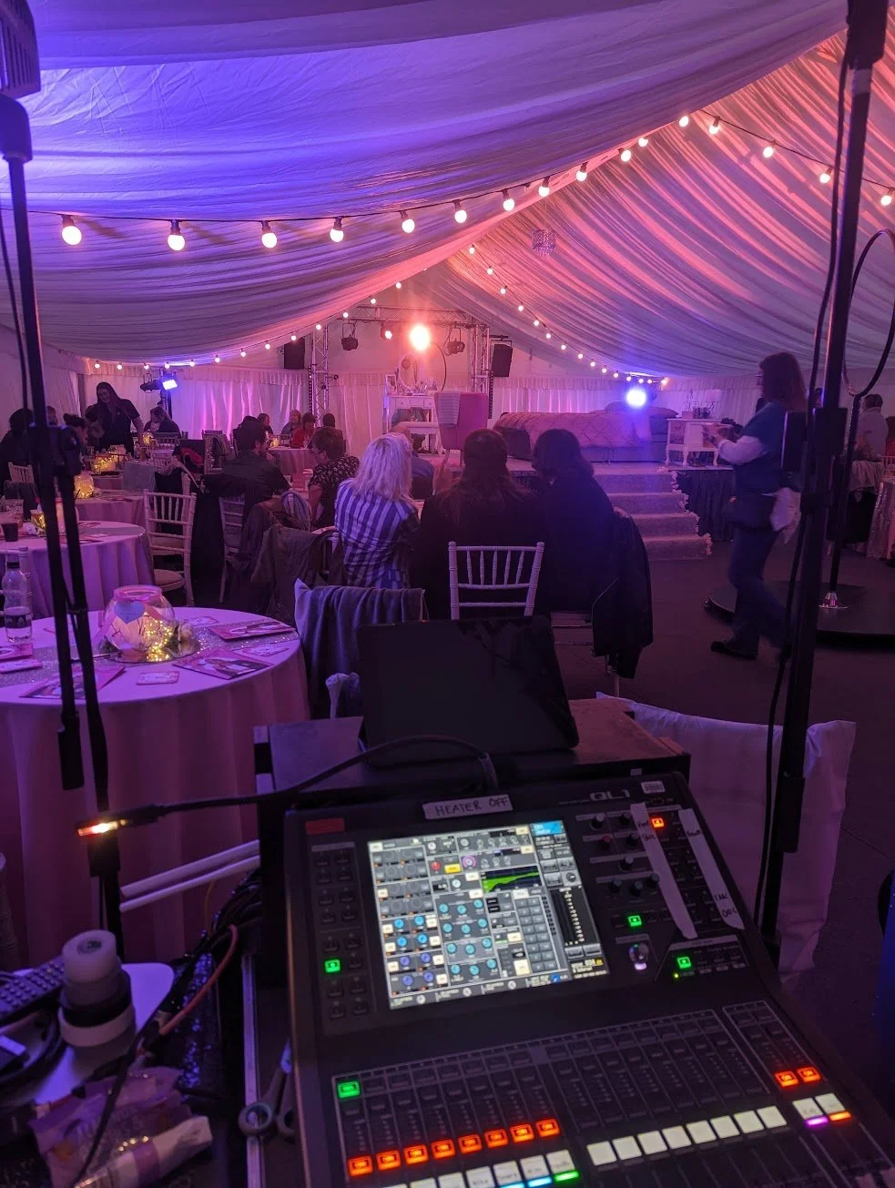 Interior of a decorated event tent with string lights, a stage with a chair and table, and guests seated at round tables. There is a soundboard in the foreground.