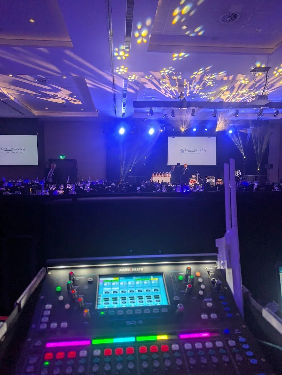 A conference or event hall with a stage, large screens displaying 'East Riding,' and an elaborate lighting setup. In the foreground, a sound mixing console with illuminated controls and a digital display.