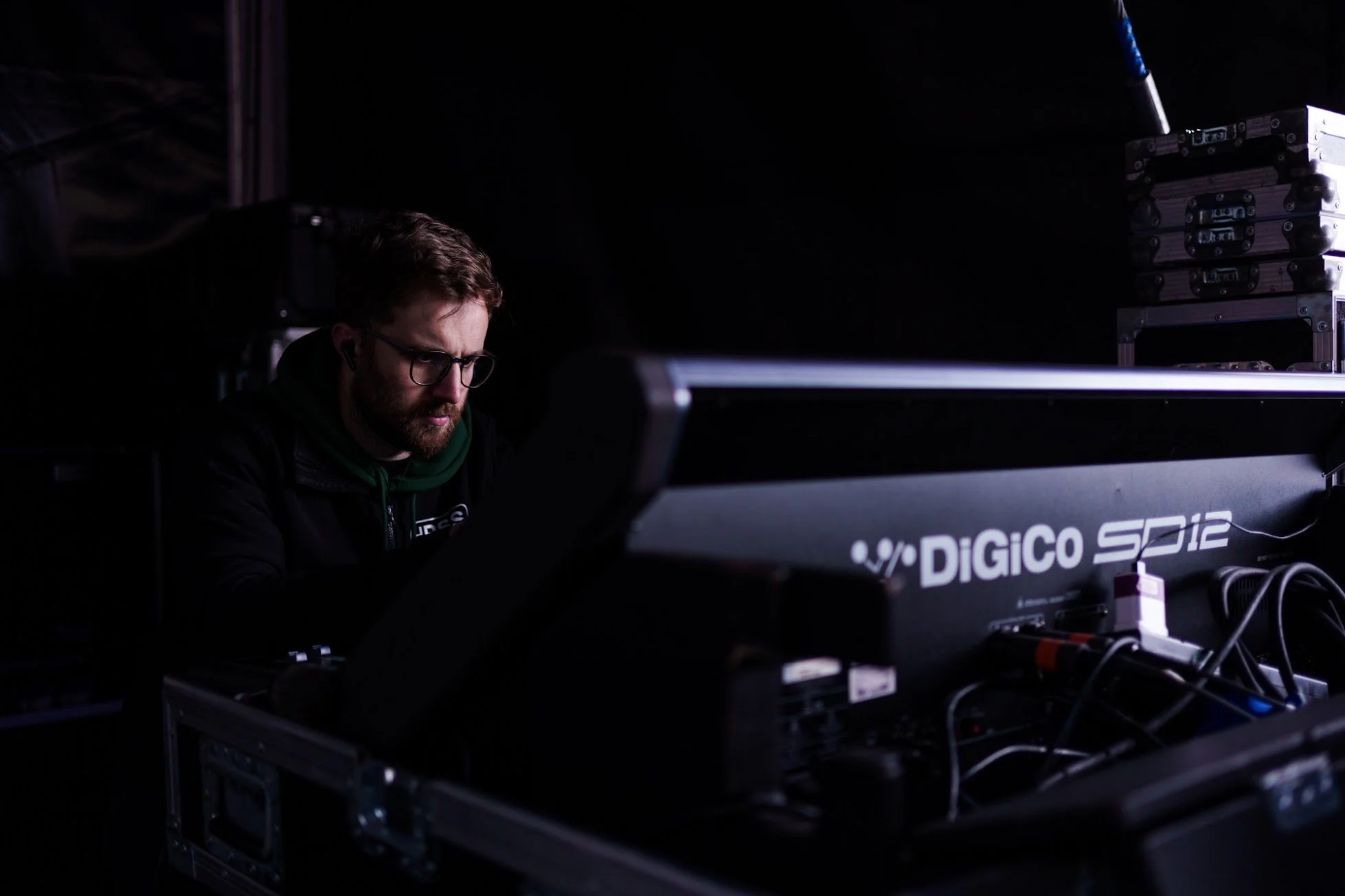 A man with glasses, beard, and dark clothing working on electronic equipment in a dark environment.