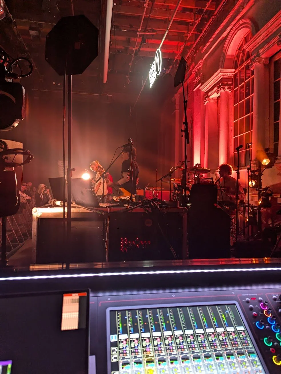 Stage with musicians performing, bright lighting, and an audience in the background, viewed from behind a mixing console.