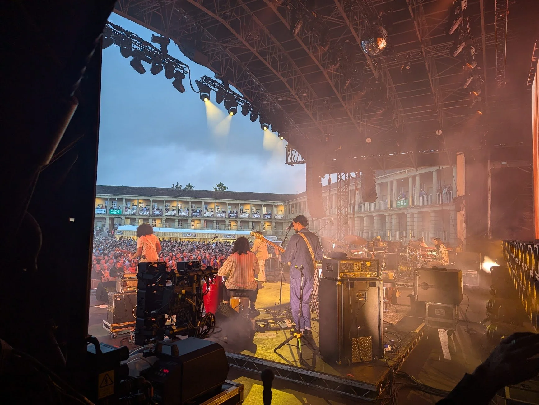 View from behind the stage of a band performing at an outdoor concert with a large audience, hotel building in the background, stage lights, and sound equipment.