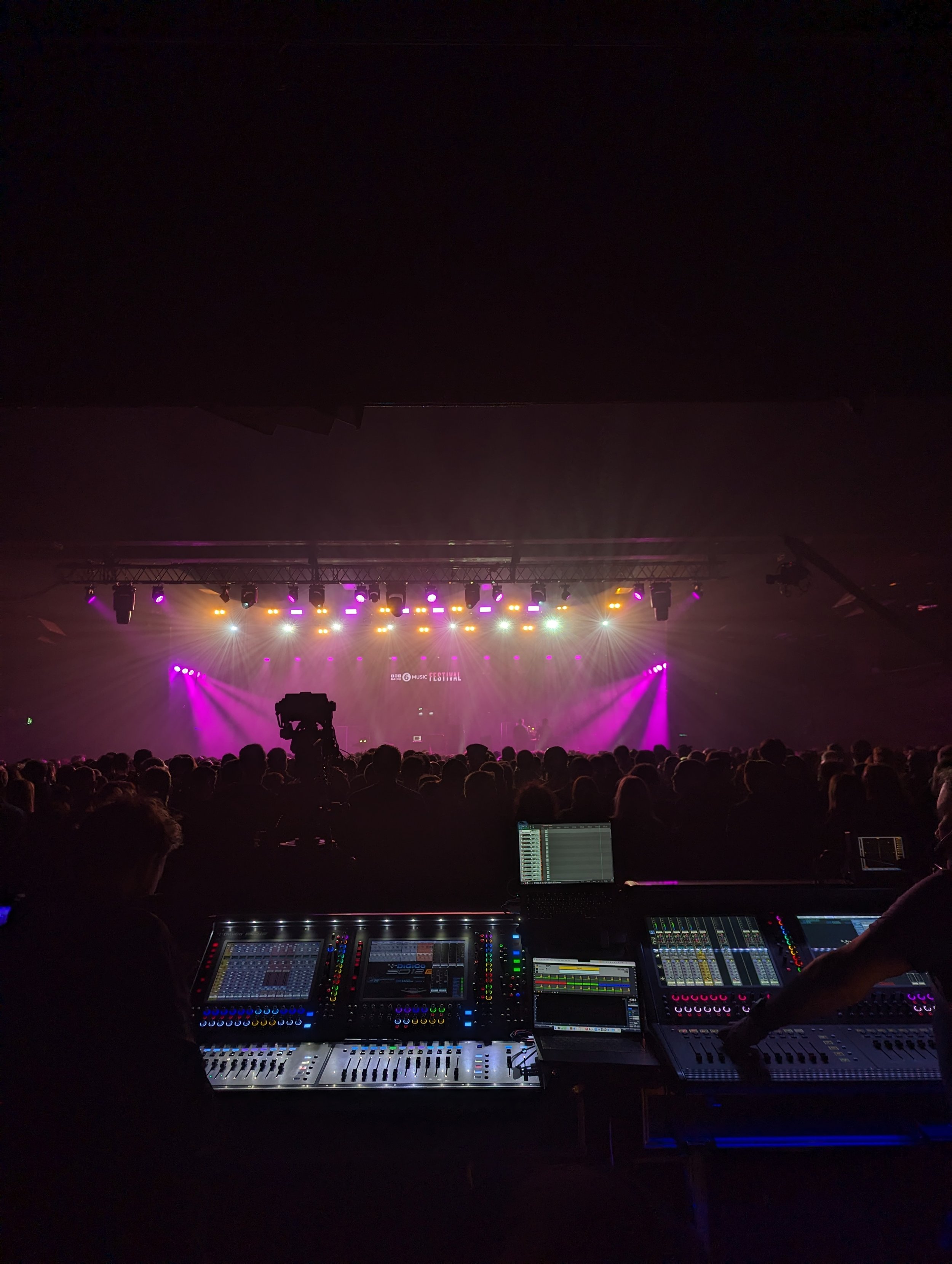 Concert stage with purple and yellow lighting, crowd facing the stage, and a sound engineer working at a mixing console in the foreground.