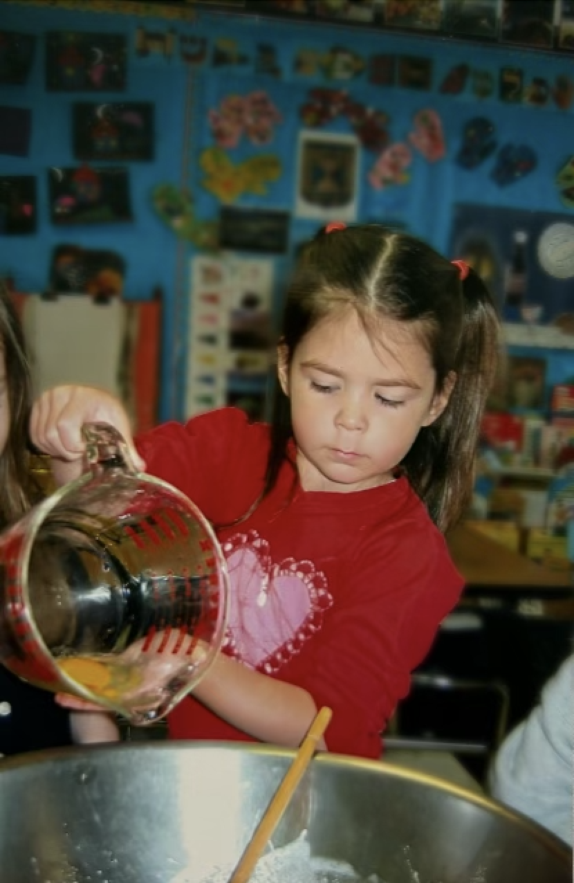 A young girl in a red shirt with a pink heart design is pouring ingredients into a mixing bowl, baking in a classroom setting.