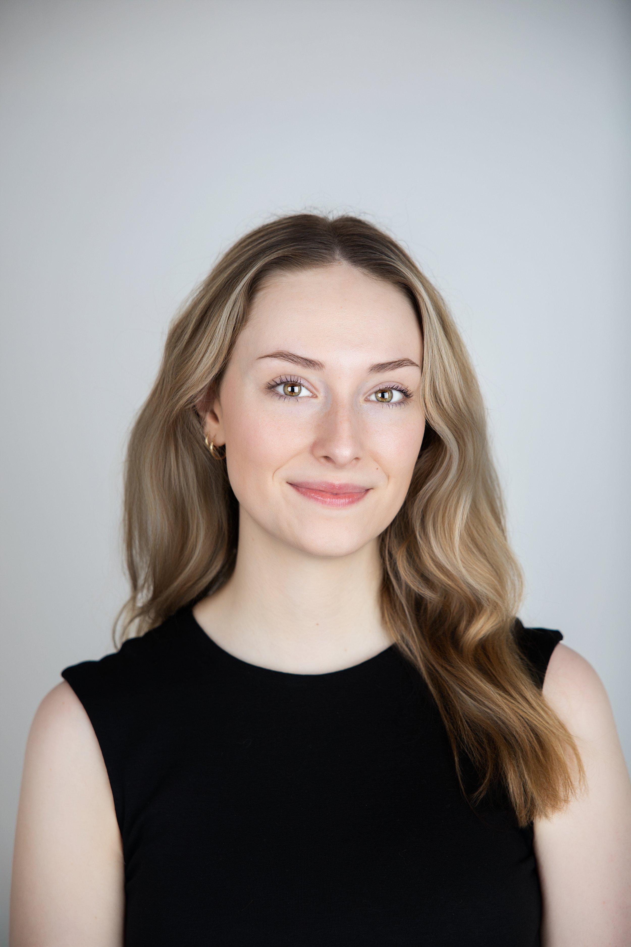 A young woman with long, wavy blonde hair, light skin, and blue eyes, wearing a black sleeveless top, smiling softly against a plain light gray background.